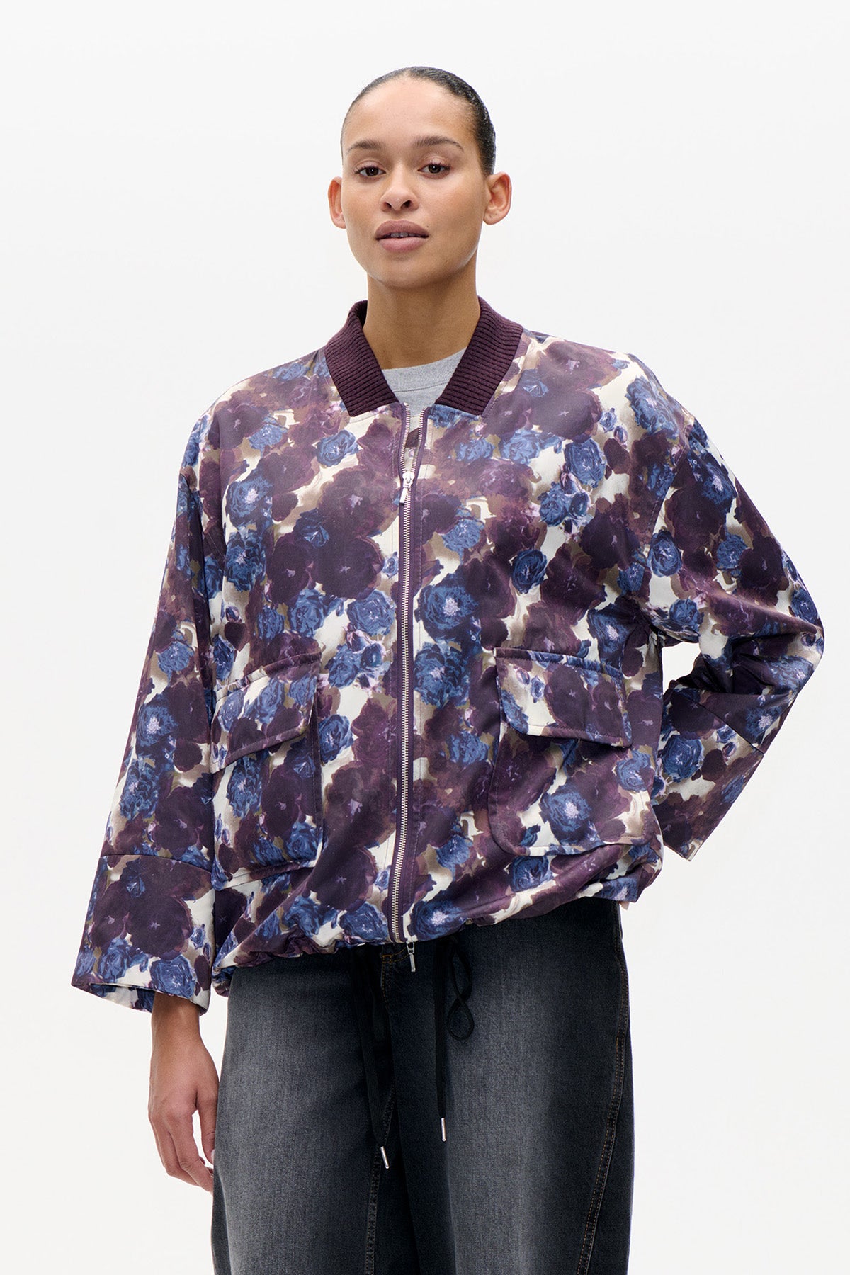 Front view of a zip-front bomber jacket with a dark plum ribbed collar, oversized blue and purple floral print, and flap pockets.