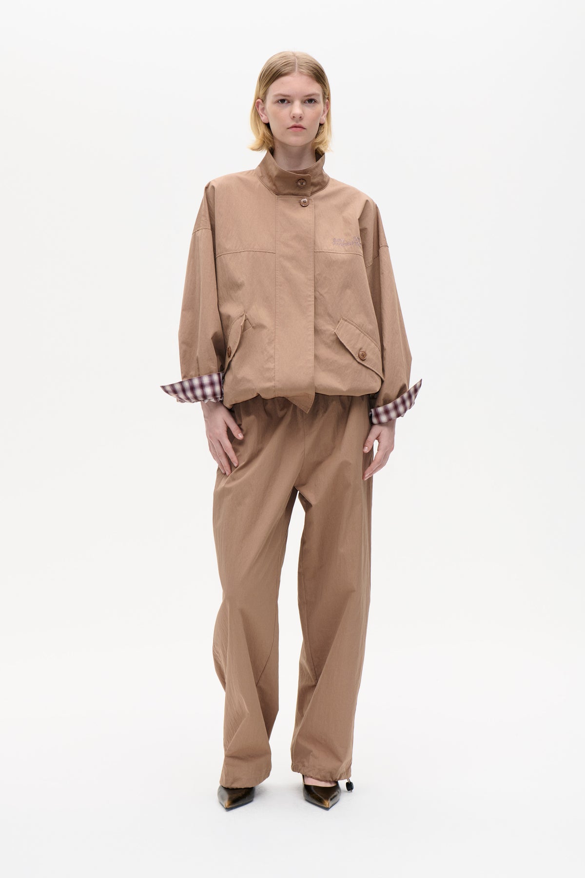 Model wearing a taupe button-front jacket with a high collar, angled pockets and plaid cuffs, paired with loose brown trousers.