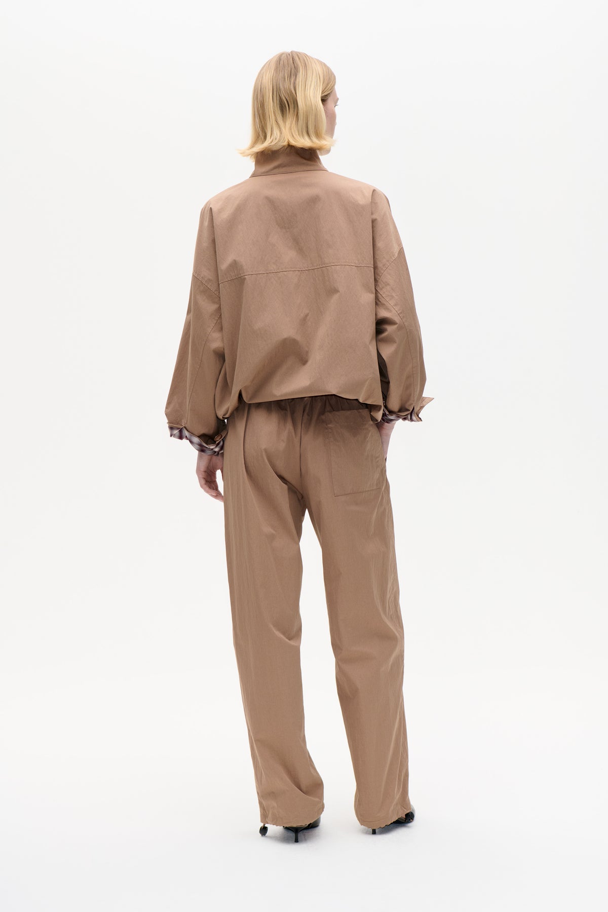 Back view of a model wearing an oversized taupe-brown collared shirt with plaid cuffs and matching wide-leg trousers.