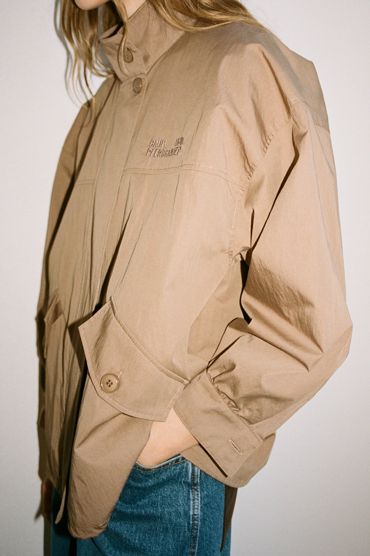Close-up campaign view of a model wearing a tan button-front jacket with stitched panels and a waist belt over blue jeans.