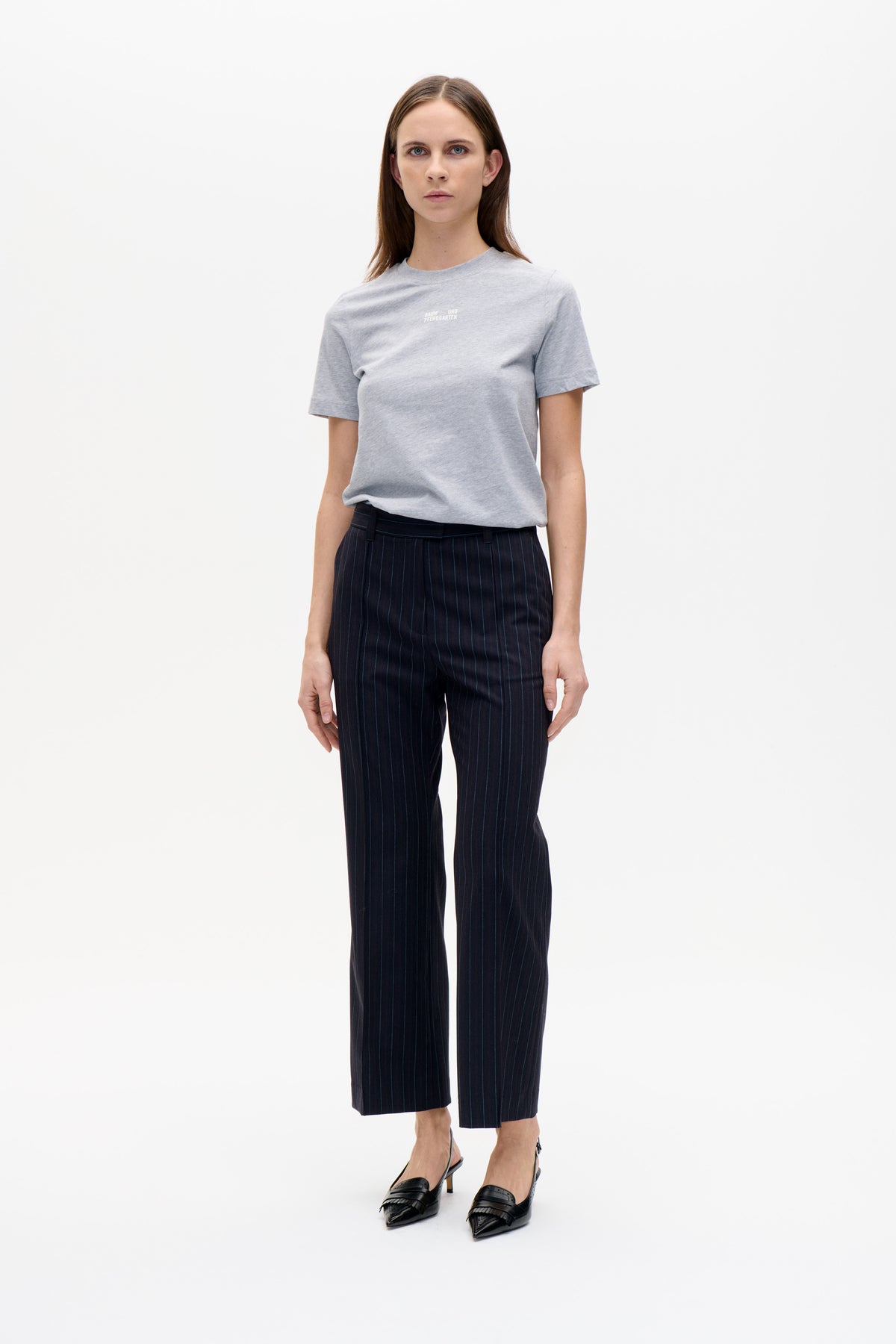 Model wearing a light grey crew-neck T-shirt with small white chest lettering and dark pinstriped trousers.