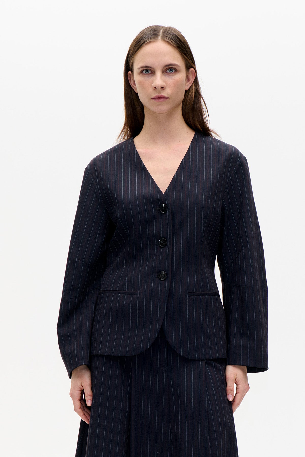 Front view of a model wearing a dark navy pinstripe blazer with a deep V-neck, three buttons and welt pockets, paired with a matching skirt.