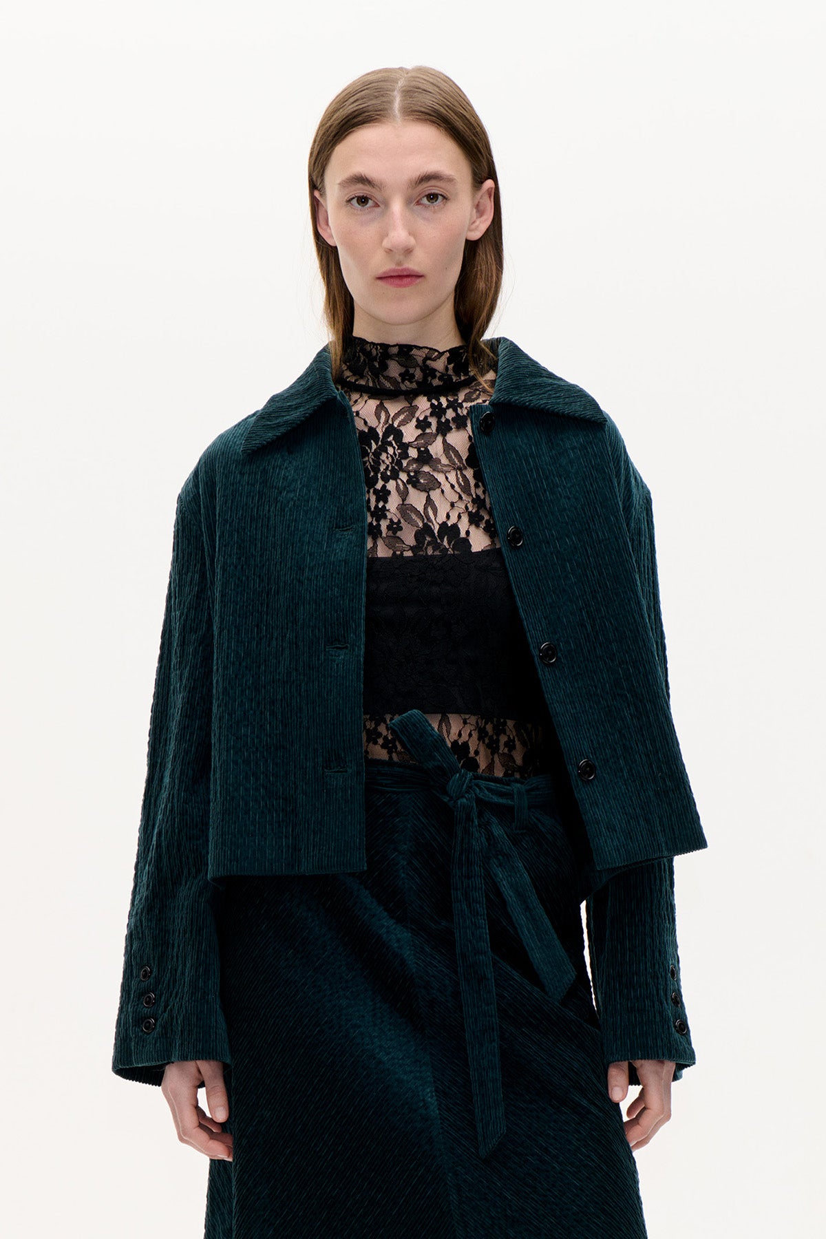 Front view of a model wearing a dark teal corduroy jacket over a black floral lace top, with a matching tie-waist skirt.