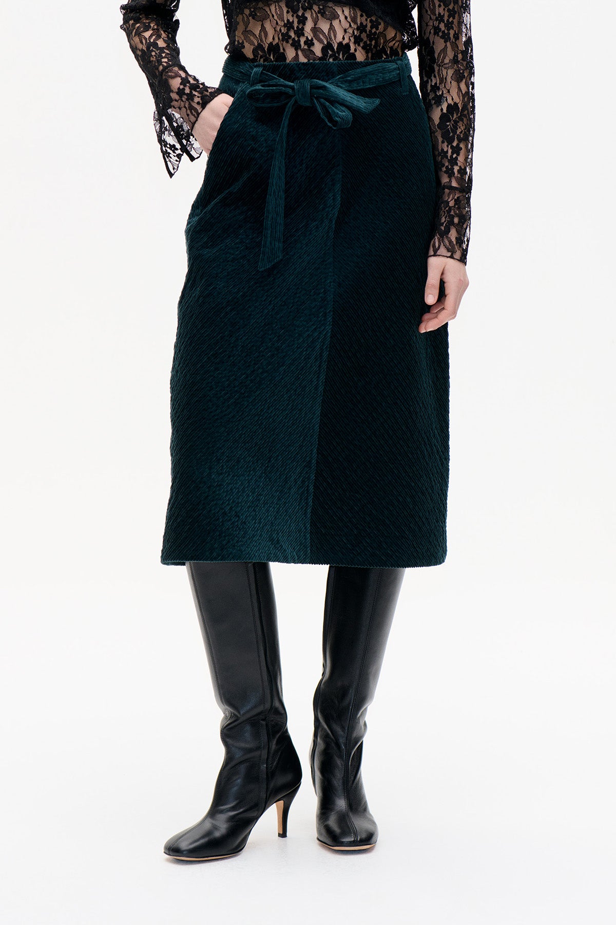 Front view of a dark teal corduroy midi skirt with a tied waist belt, styled with a black lace top and knee-high boots.
