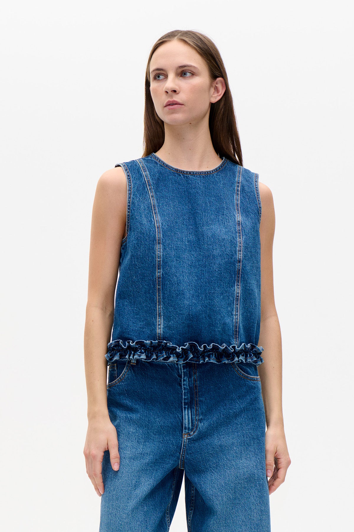Front view of a sleeveless blue denim top with contrast stitching and a ruffled hem, worn with matching jeans.