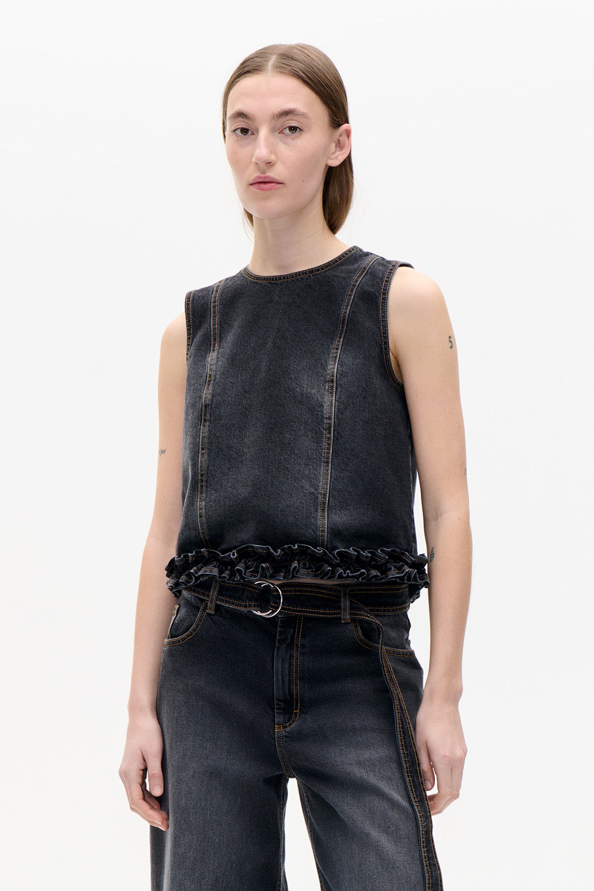 Model wearing a charcoal washed denim sleeveless top with contrast stitching and a ruffled hem, paired with matching jeans.