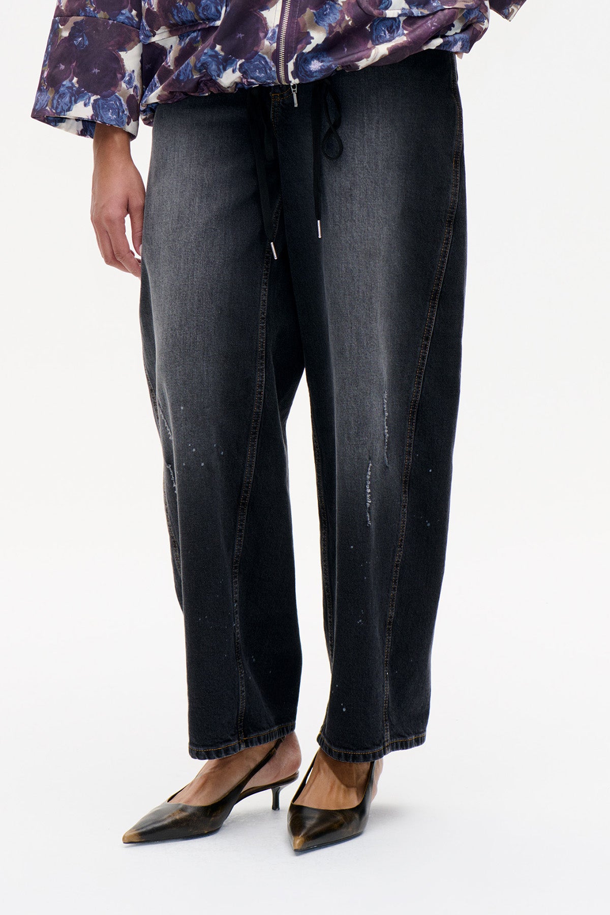 Detail van de voorkant van antracietzwarte jeans met trekkoord in de taille, rechte pijpen en verspreide slijtage-accenten, gestyled met een bloemenjas.