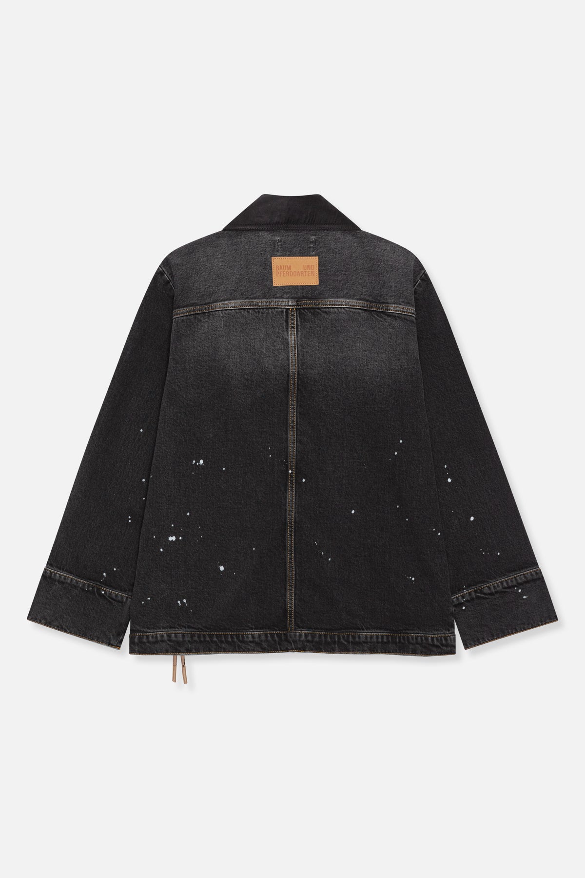 Back view of a charcoal-black denim jacket with contrast stitching, a tan collar patch and scattered white paint-like spots.