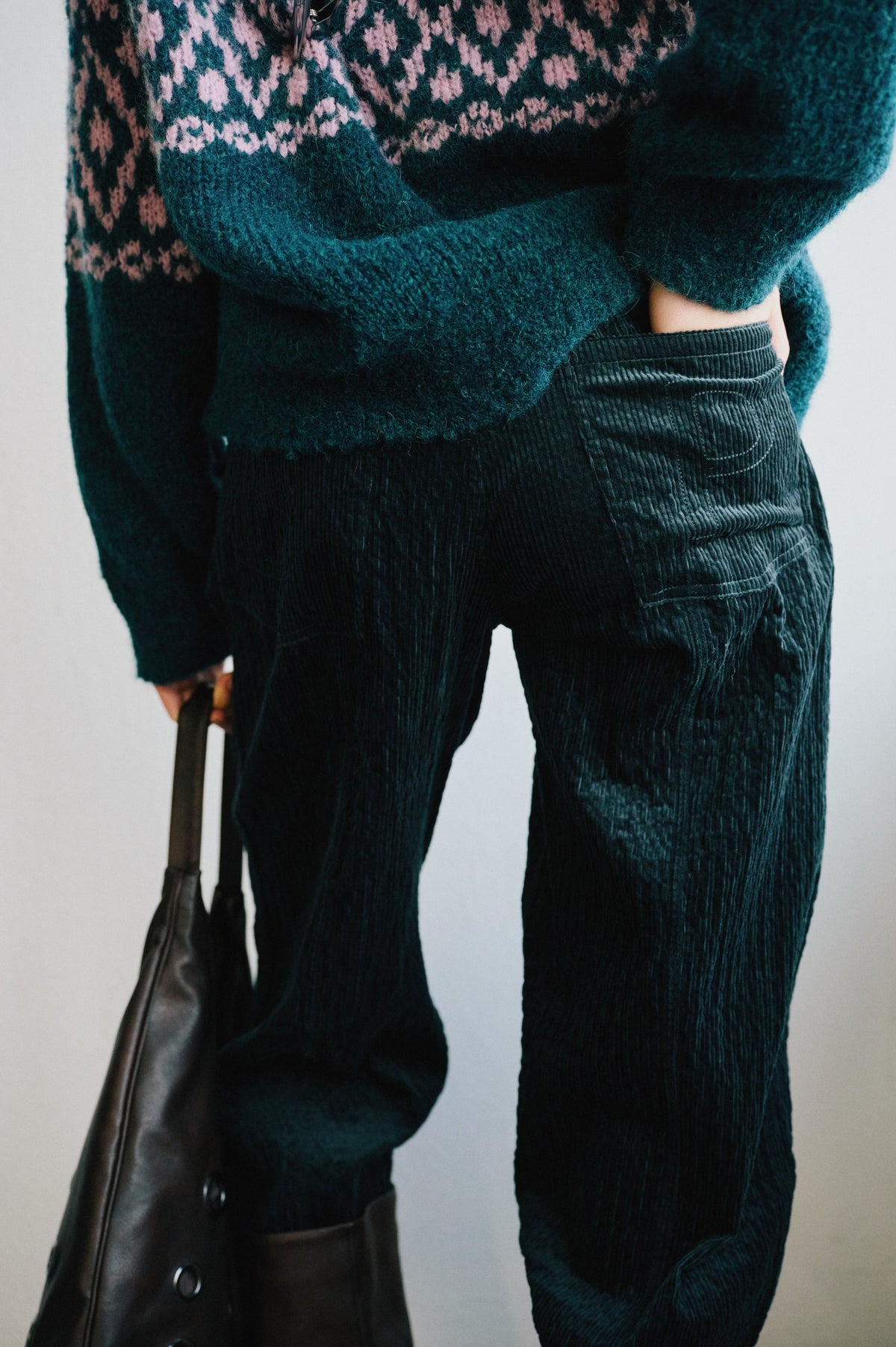Close-up of a teal knit cardigan with a pink pattern over dark corduroy trousers, with a black leather bag at the side