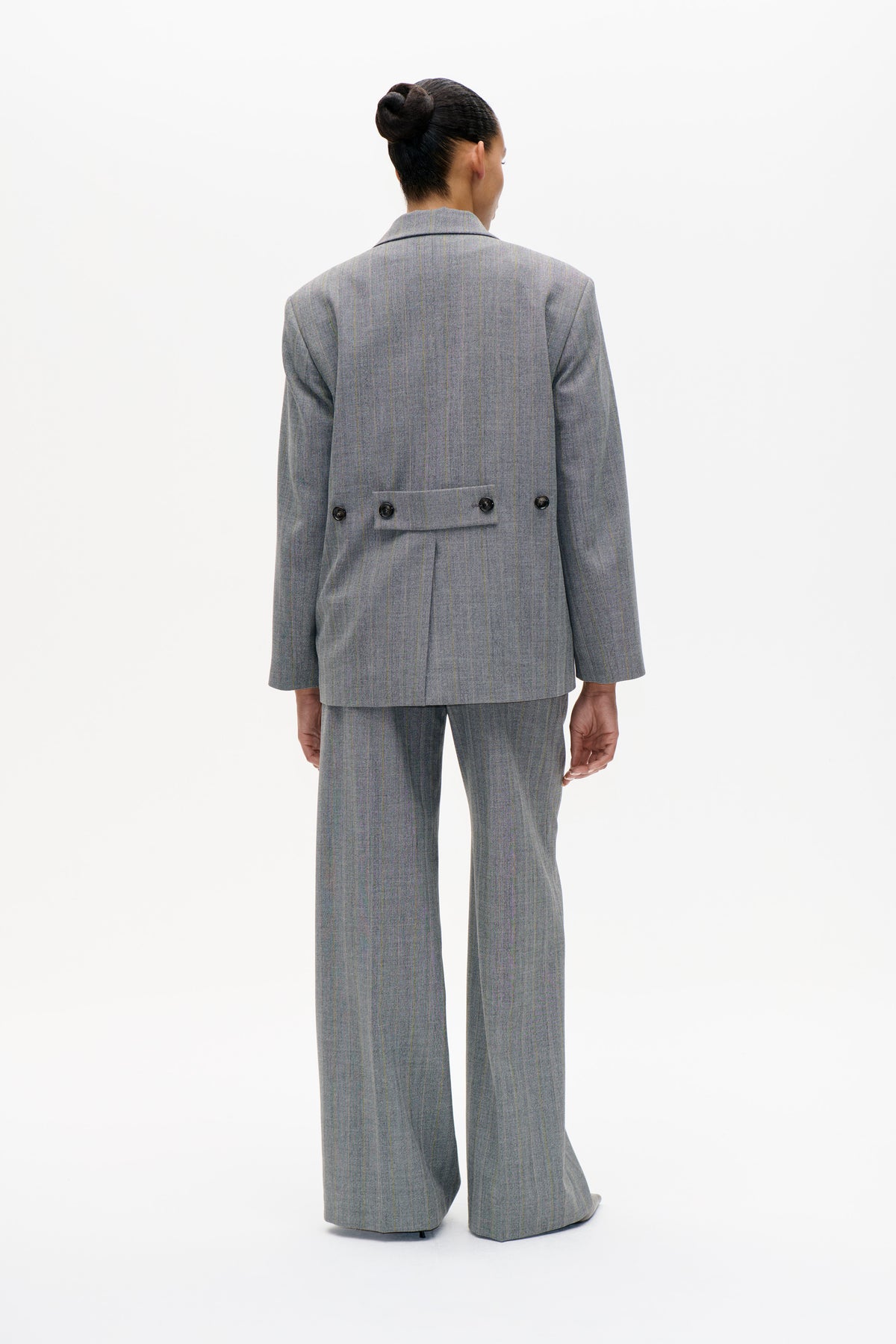 Rear view of a model wearing a grey pinstripe blazer with a buttoned back tab and matching wide-leg trousers.