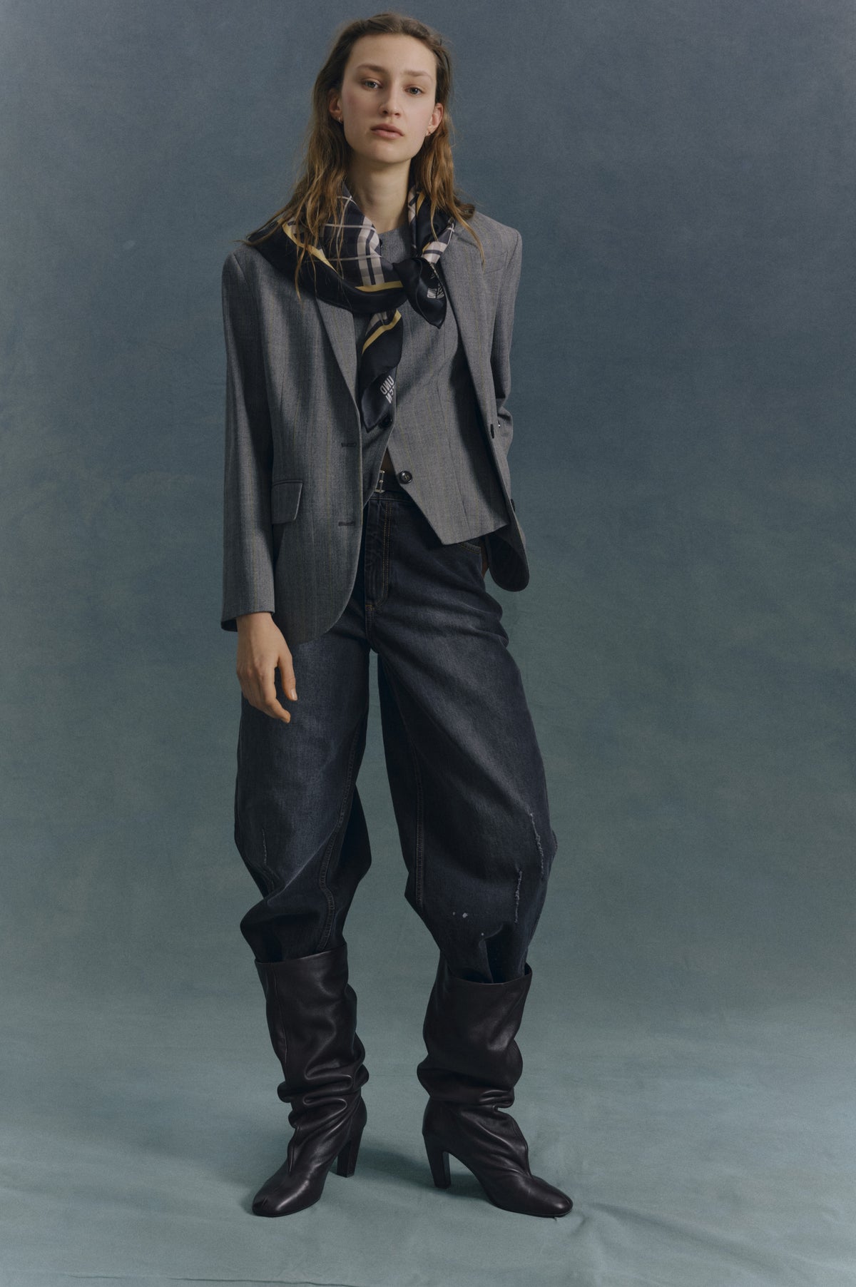 Campaign portrait of a model wearing a grey oversized blazer, dark wide-leg jeans, black knee-high boots and a navy checked scarf against a blue-grey backdrop.