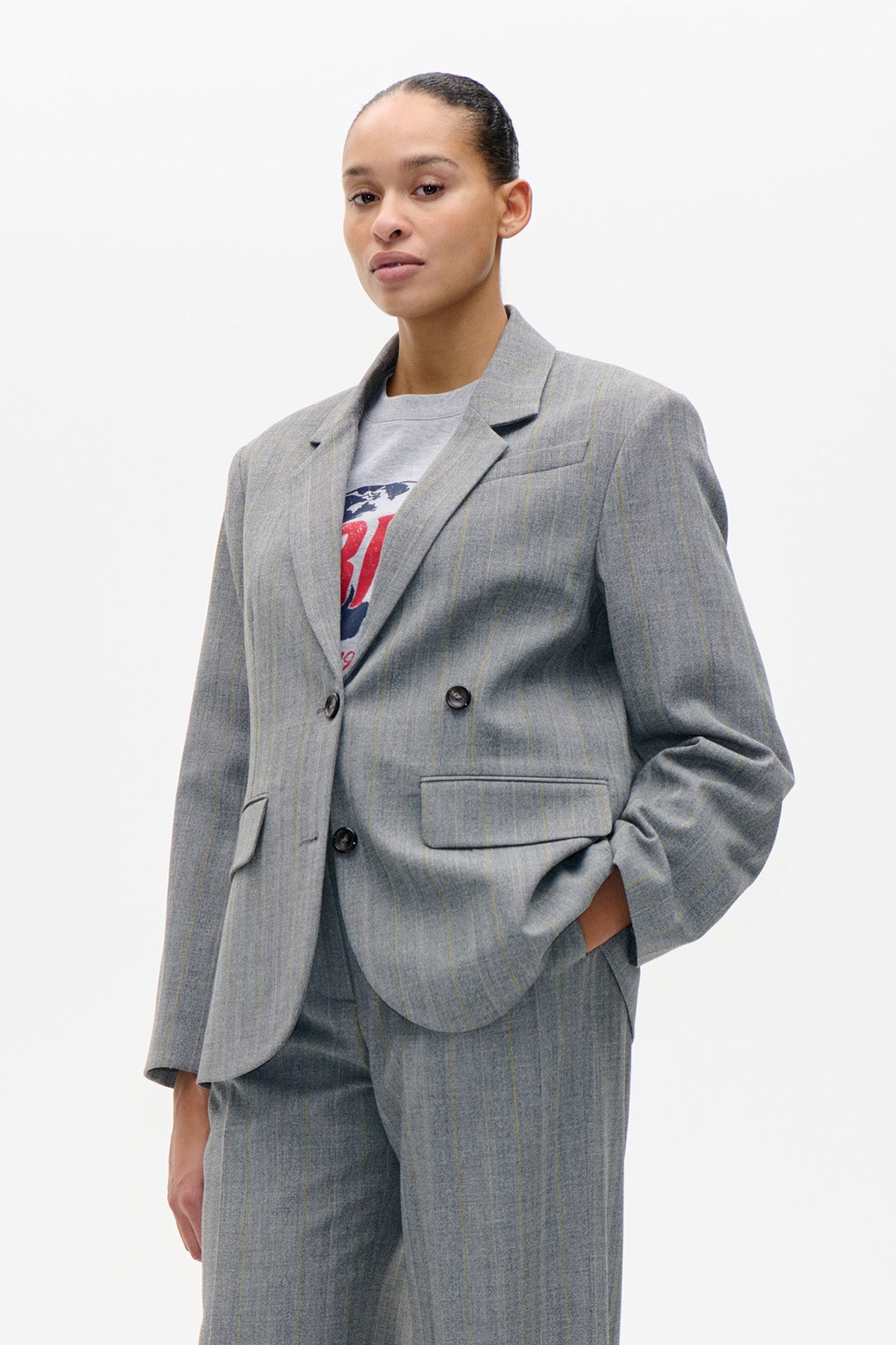 Front view of a grey pinstripe double-breasted blazer with notched lapels and flap pockets, styled with matching trousers and a graphic T-shirt.