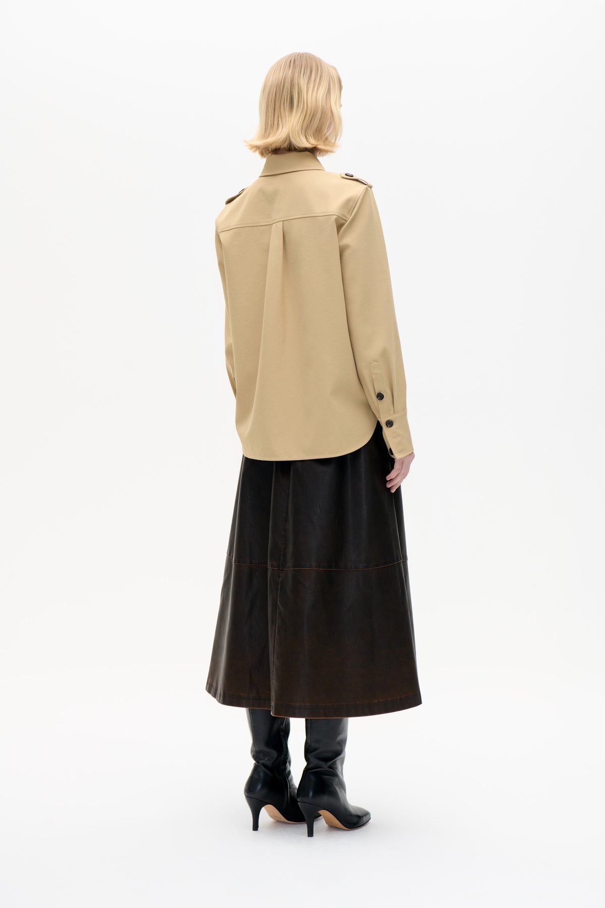 Back view of a beige shirt jacket with shoulder tabs and a layered back yoke, styled with a black skirt and heeled boots.