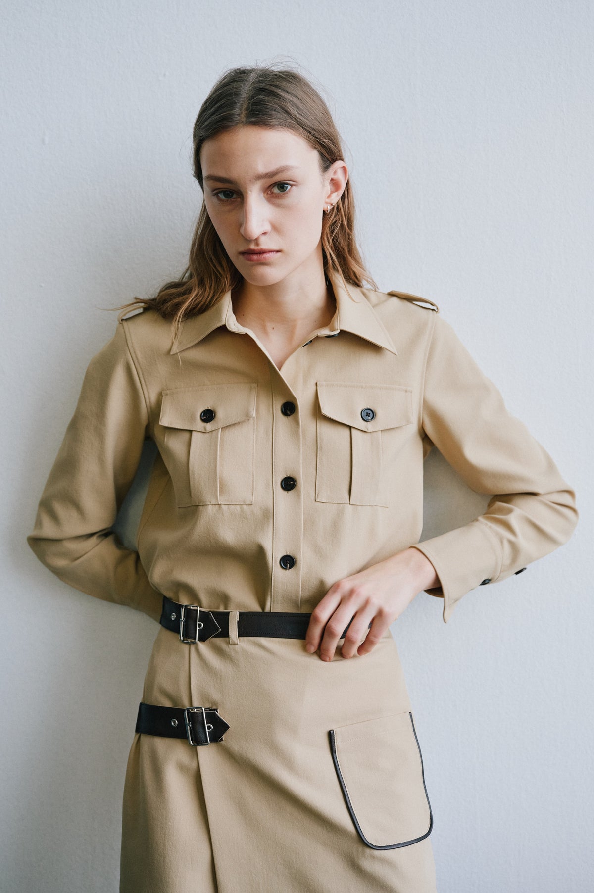 Campaign portrait showing the front of a beige belted shirt dress with black buttons, chest pockets and dark-trimmed flap pocket.