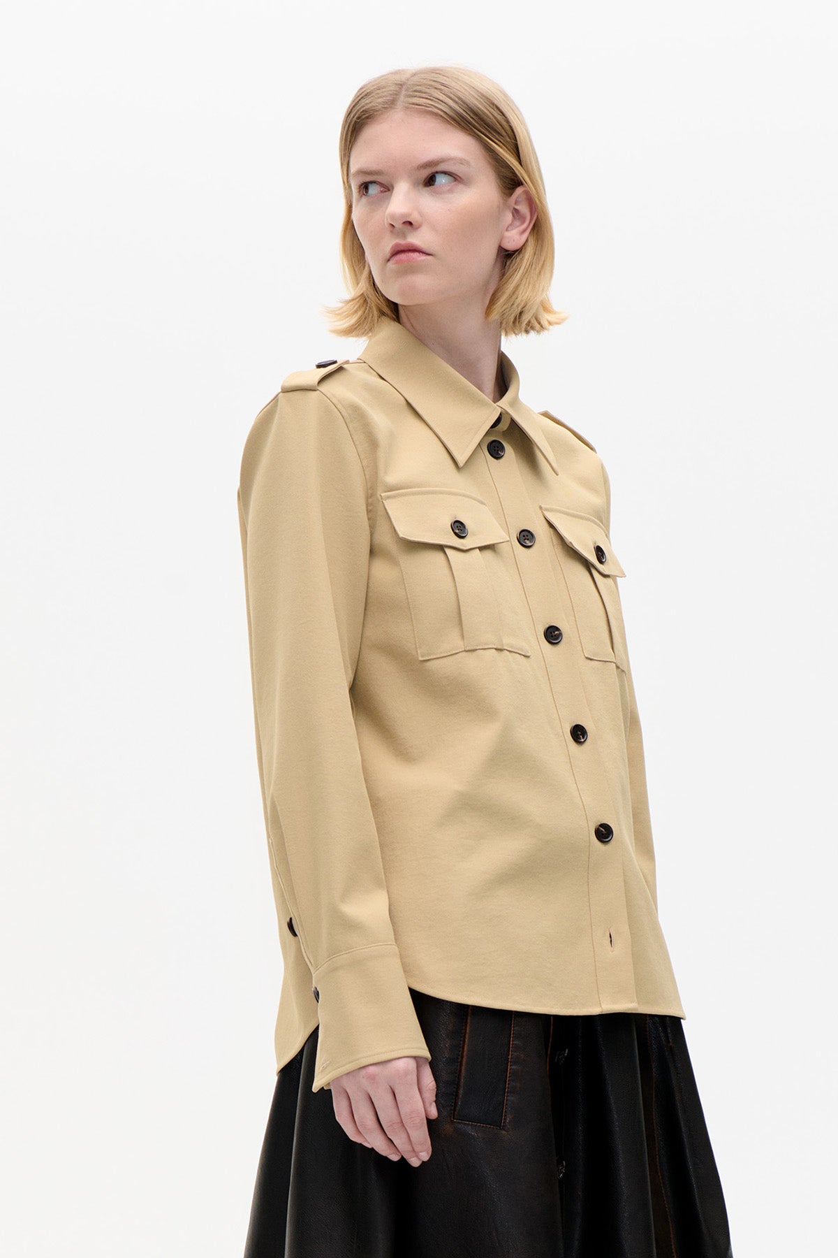 Model wearing a beige button-front shirt jacket with a pointed collar, shoulder tabs and flap chest pockets, paired with a black skirt.
