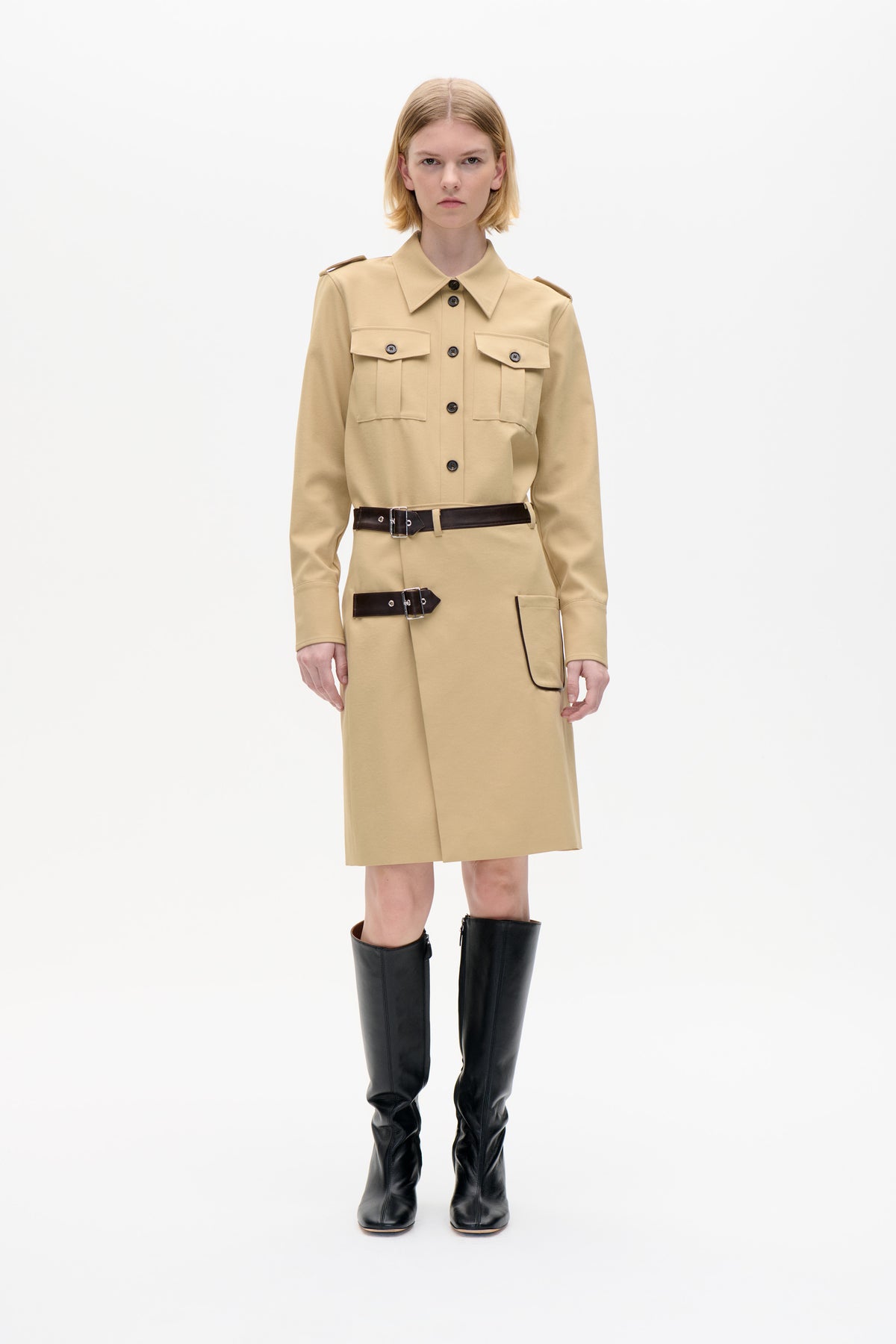Front view of a sand-beige shirt dress with pointed collar, button front, flap chest pockets and dark buckle straps, styled with black knee-high boots.