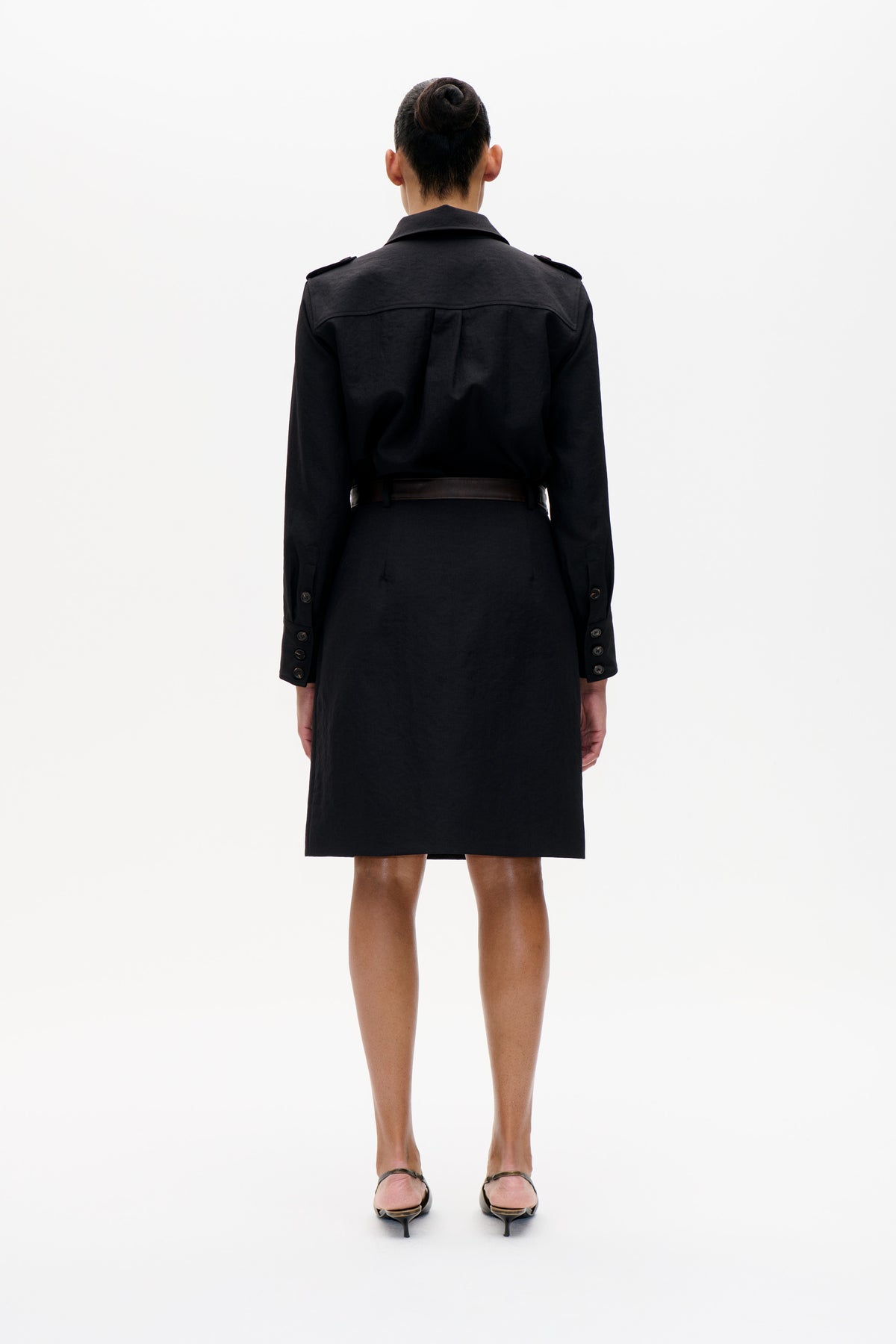 Back view of a model wearing a black knee-length coat with shoulder tabs, a cape-like back yoke, buttoned cuffs and a dark belt.