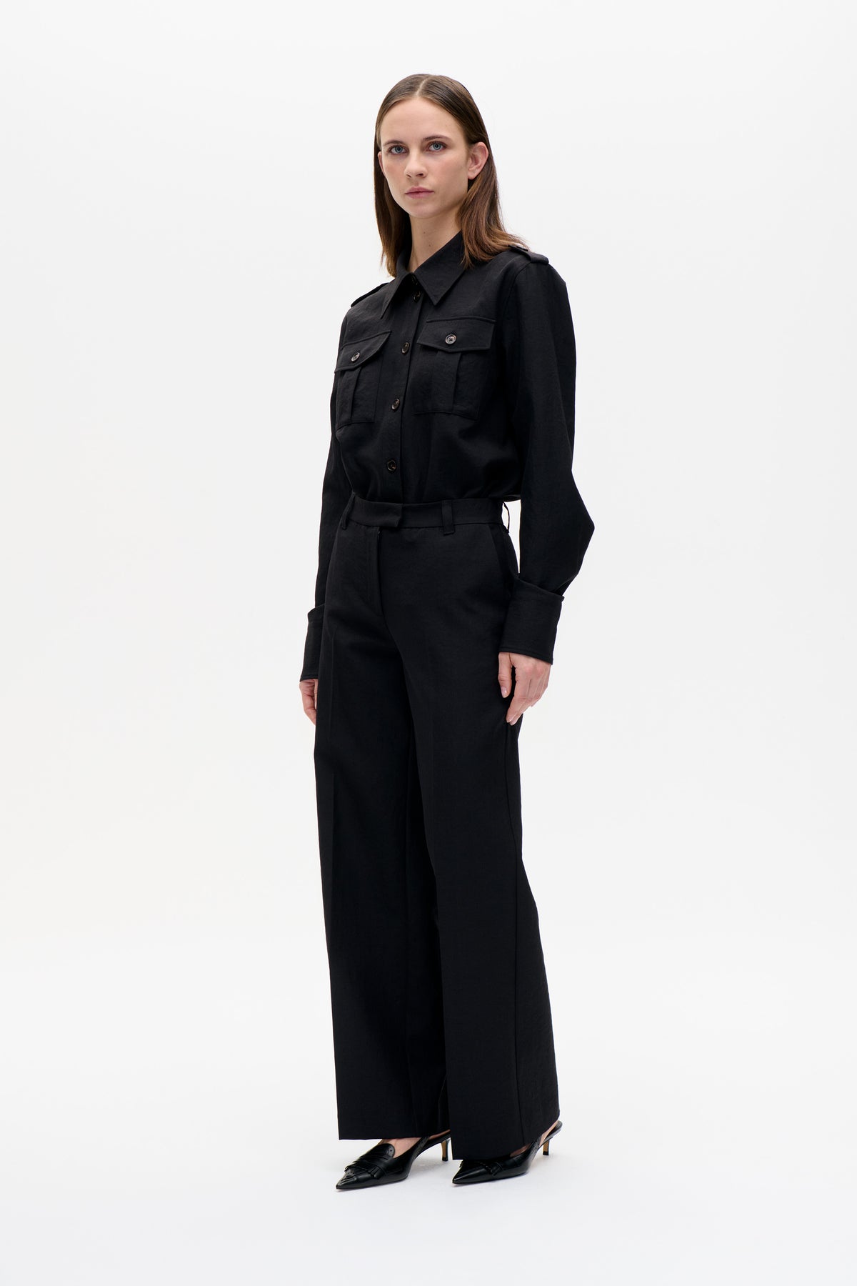 Model wearing a black button-front shirt jacket with flap chest pockets and a waist tie, paired with wide-leg trousers.