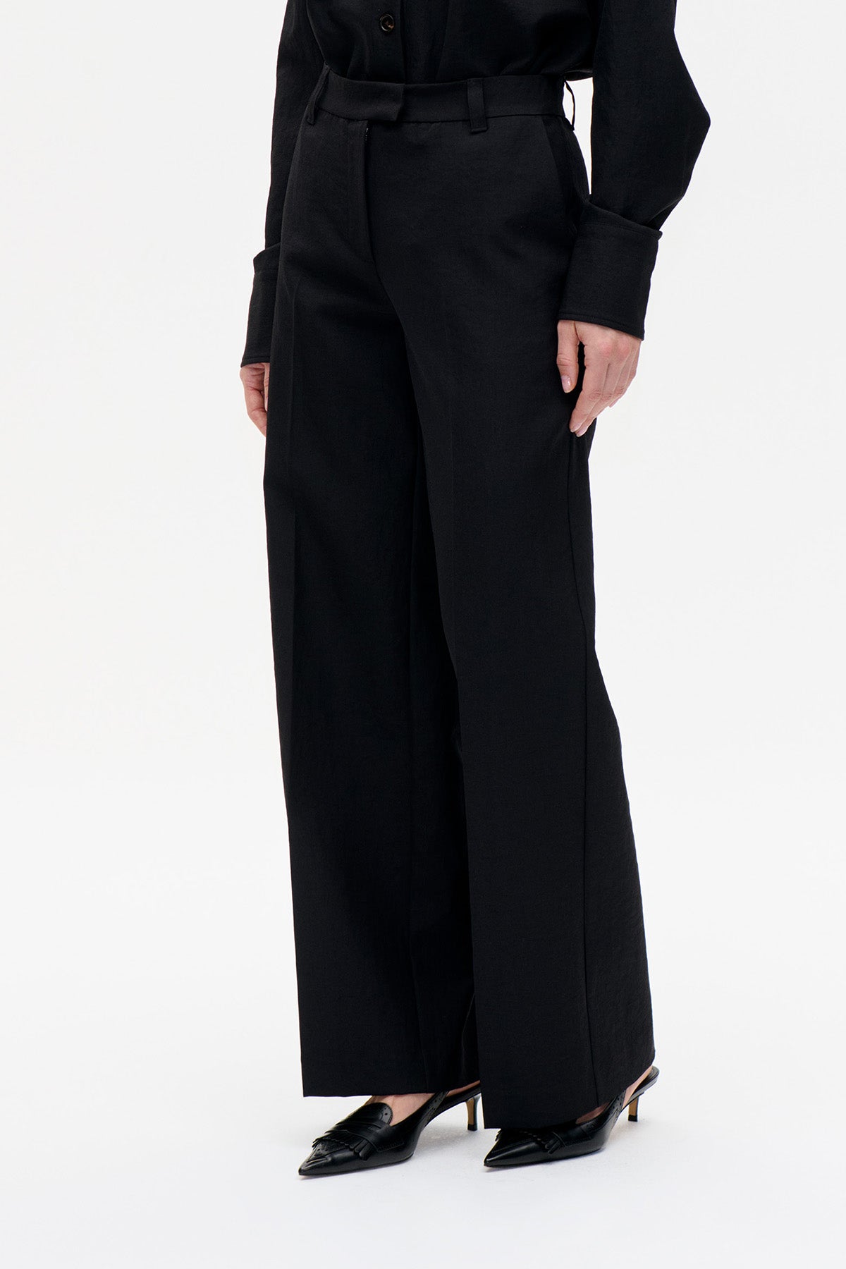 Close-up front view of black wide-leg trousers with a high waistband and belt loops, worn with a black blouse.