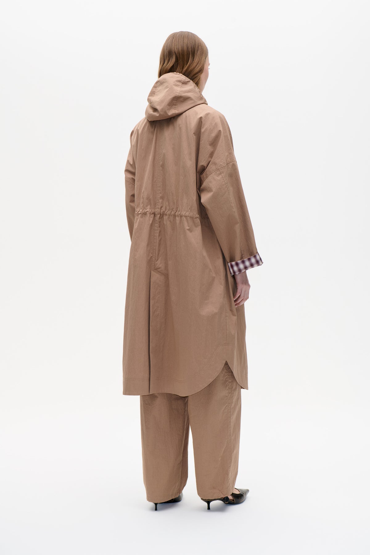 Back view of a model wearing a long taupe hooded coat with a gathered waist and burgundy plaid sleeve cuffs, paired with matching trousers.