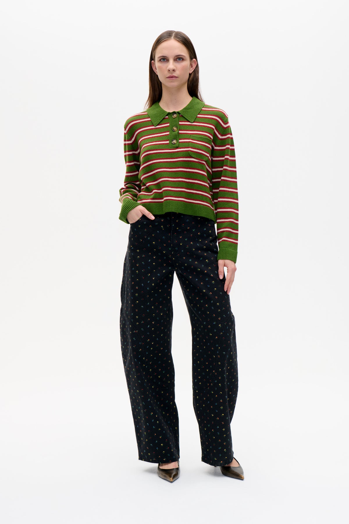 Front view of a green knit polo sweater with red and white stripes, styled with black trousers covered in tiny multicoloured motifs.