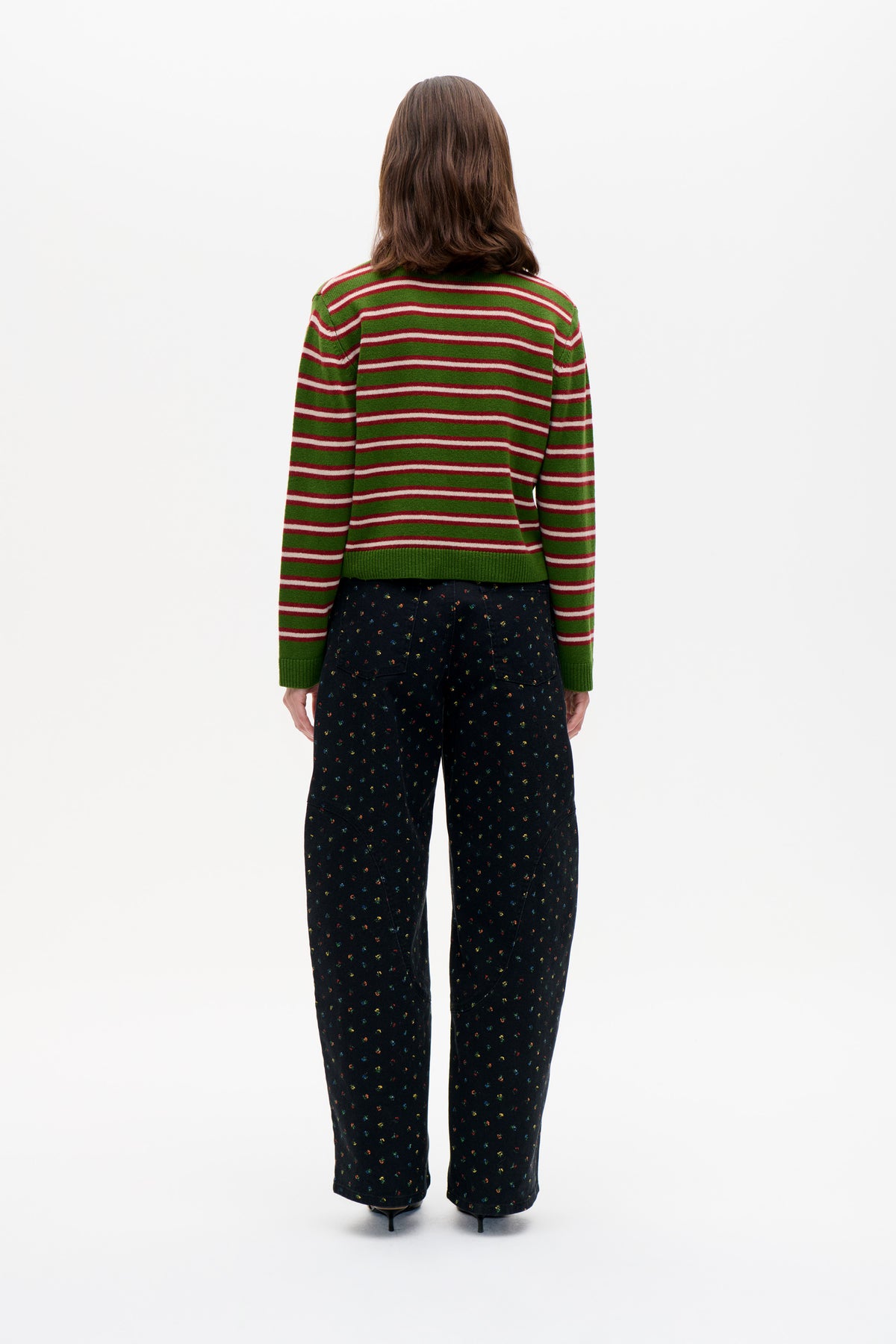Back view of a model wearing a green sweater with red and white horizontal stripes and black trousers with tiny multicoloured motifs.