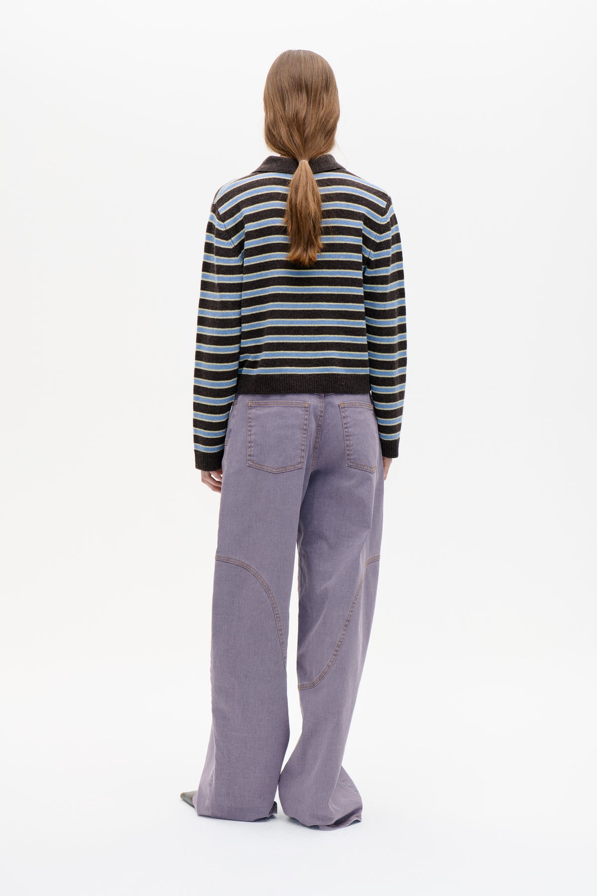 Back view of a model wearing a dark striped knit sweater with blue and pale yellow bands, paired with loose lavender trousers.
