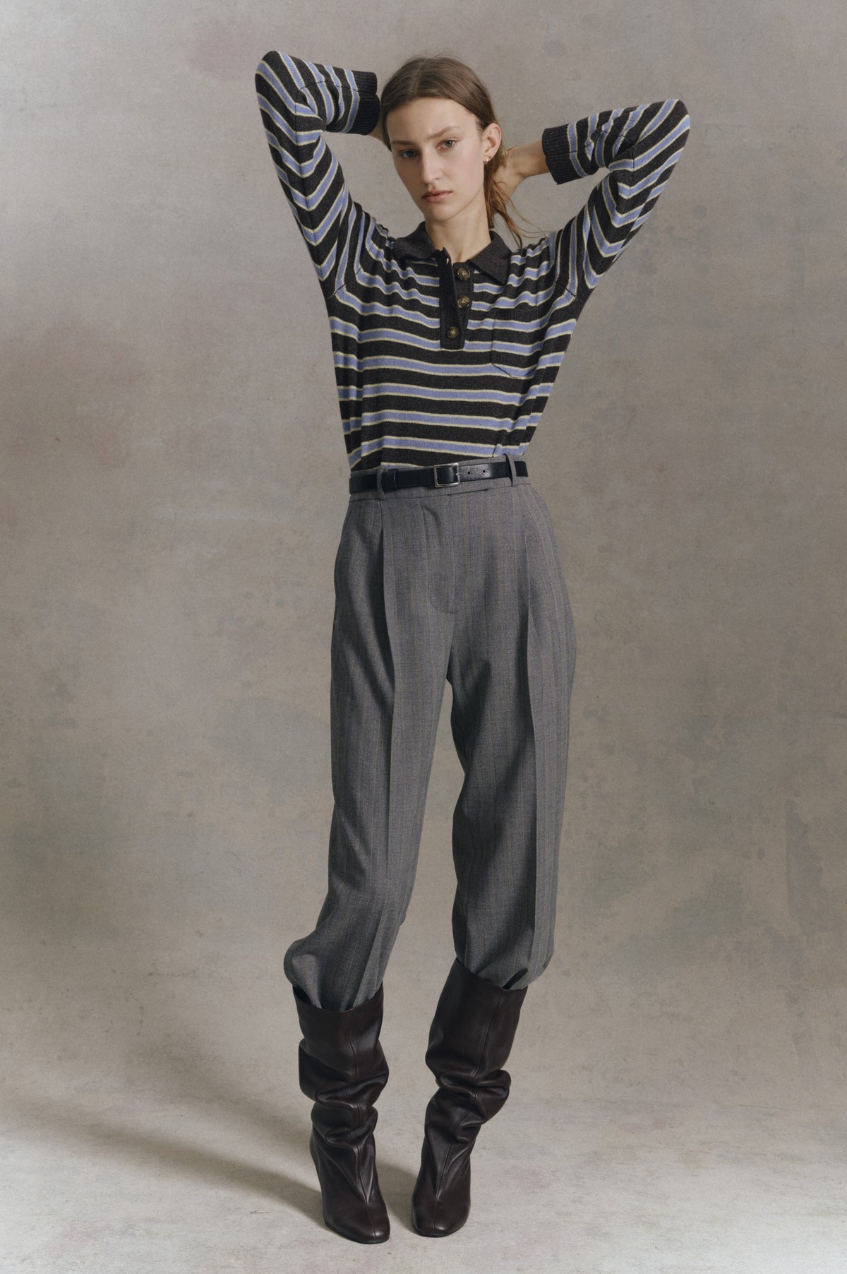 Fashion model poses against a neutral backdrop in a charcoal striped button-front knit top, grey trousers and tall dark boots.