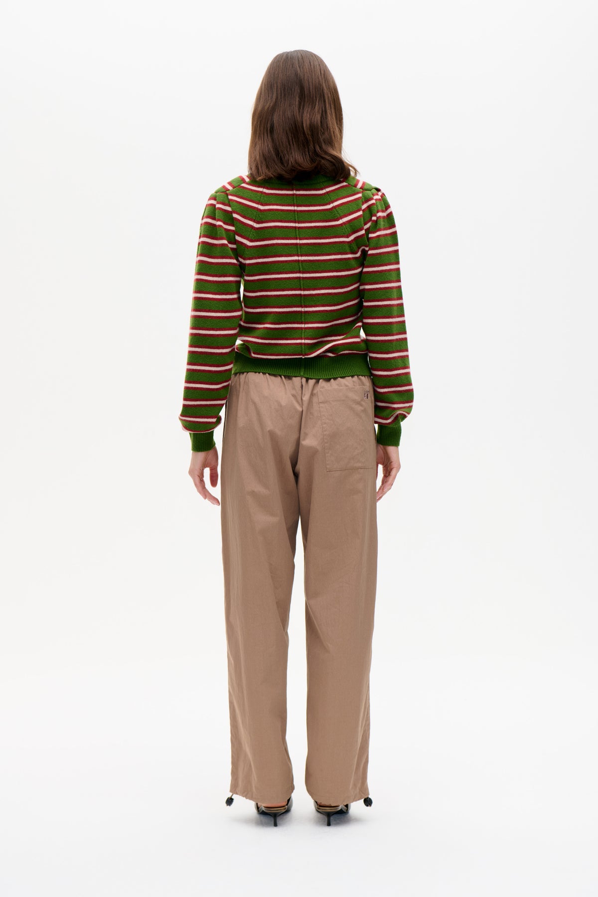 Back view of a model wearing a green knit sweater with red and white horizontal stripes and ribbed cuffs, paired with taupe wide-leg trousers.