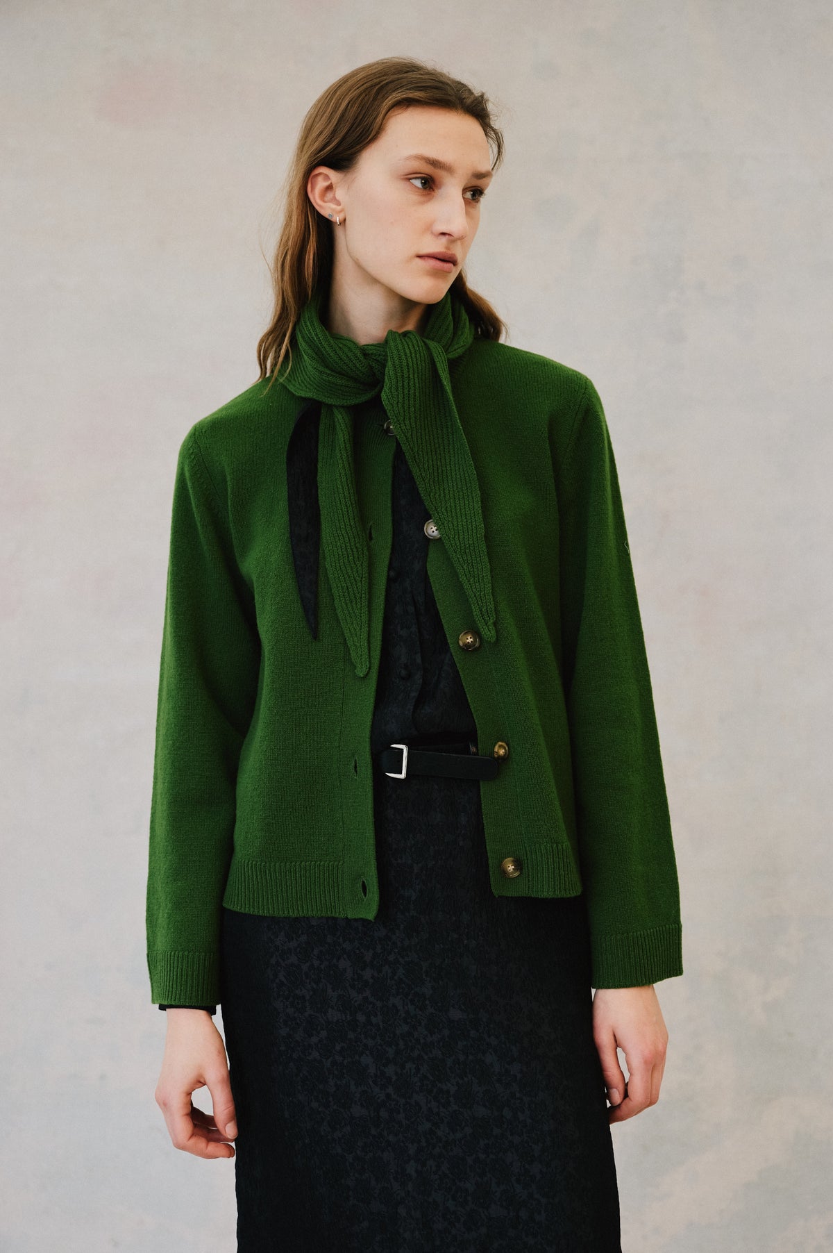 Campaign portrait of a model wearing a green button-front cardigan and matching ribbed scarf over a black patterned dress with a belt.