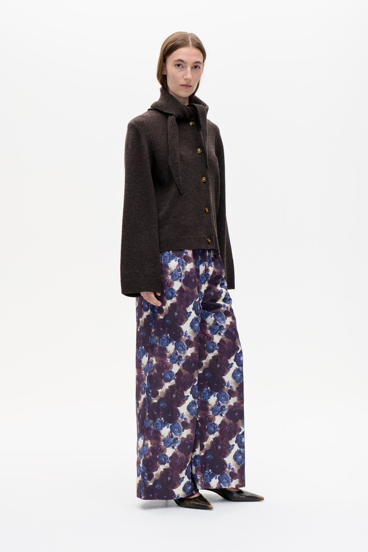 Model wearing a dark brown button-front cardigan and ribbed scarf with wide-leg cream trousers printed with blue and plum flowers.