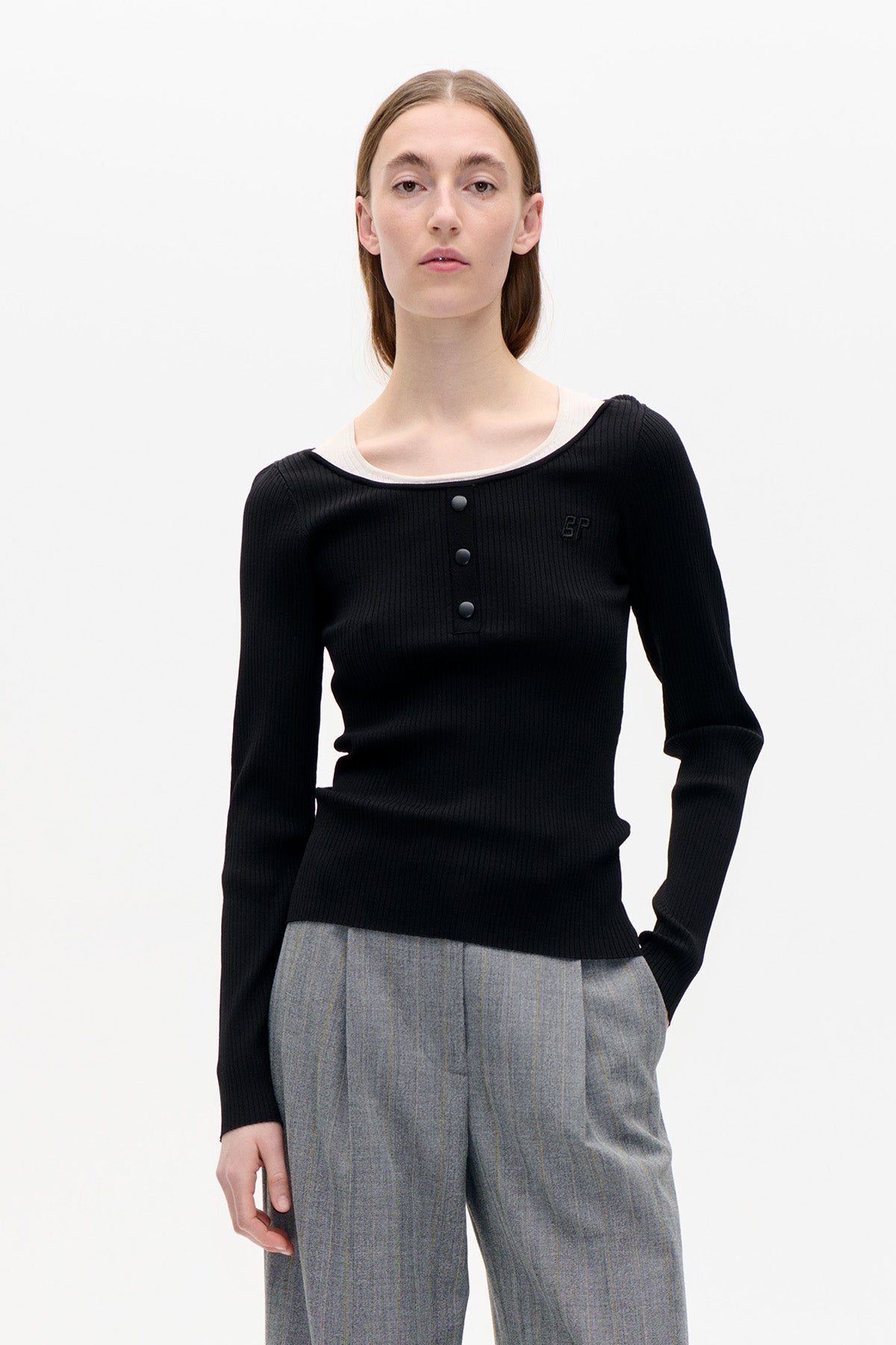 Front view of a black ribbed long-sleeve top with a wide neckline, three buttons and a small chest emblem, worn with grey pinstriped trousers.