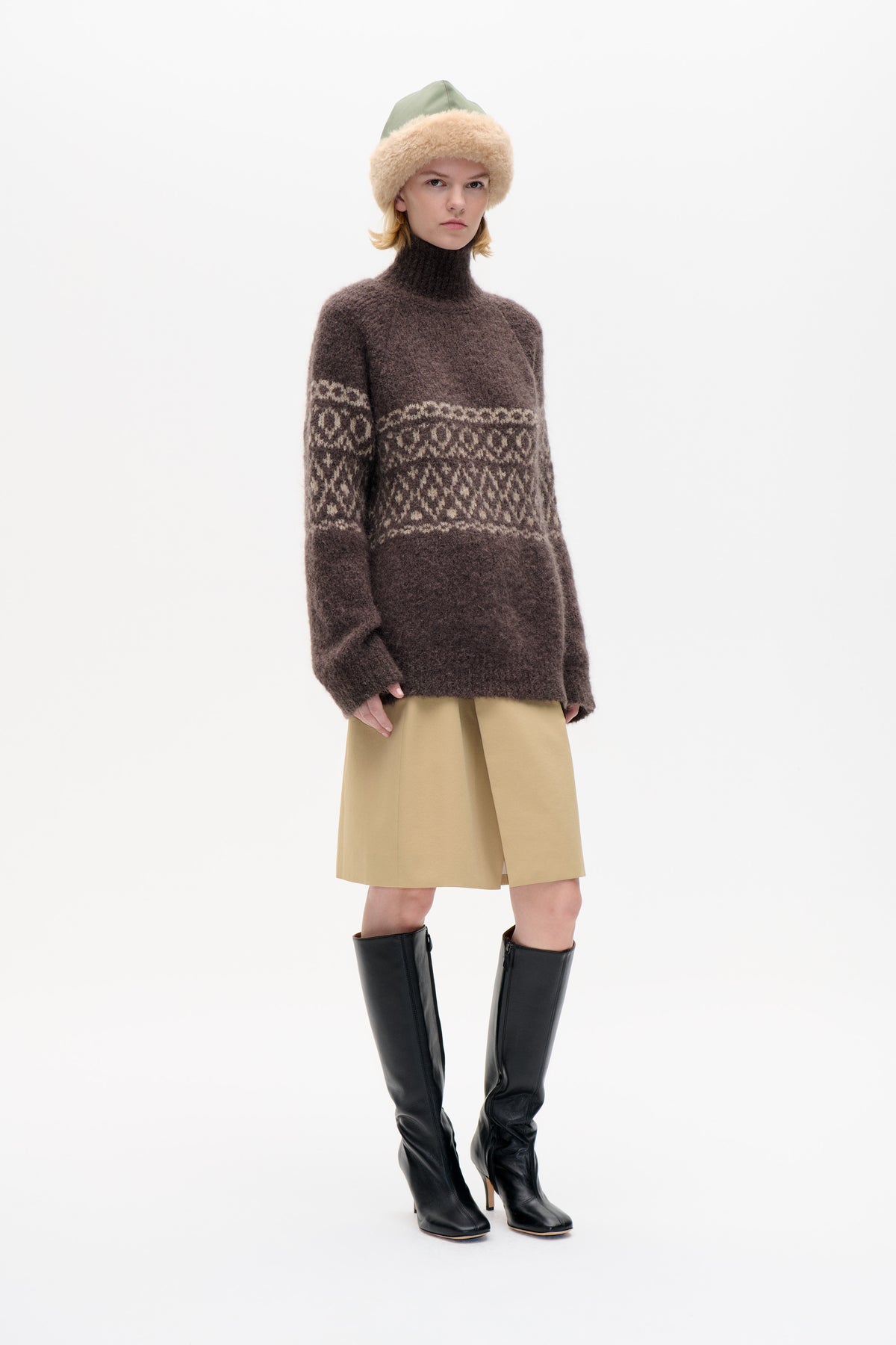 Model wearing a brown high-neck sweater with cream geometric bands, a tan skirt, a green cap with a plush beige brim, and black knee-high boots.