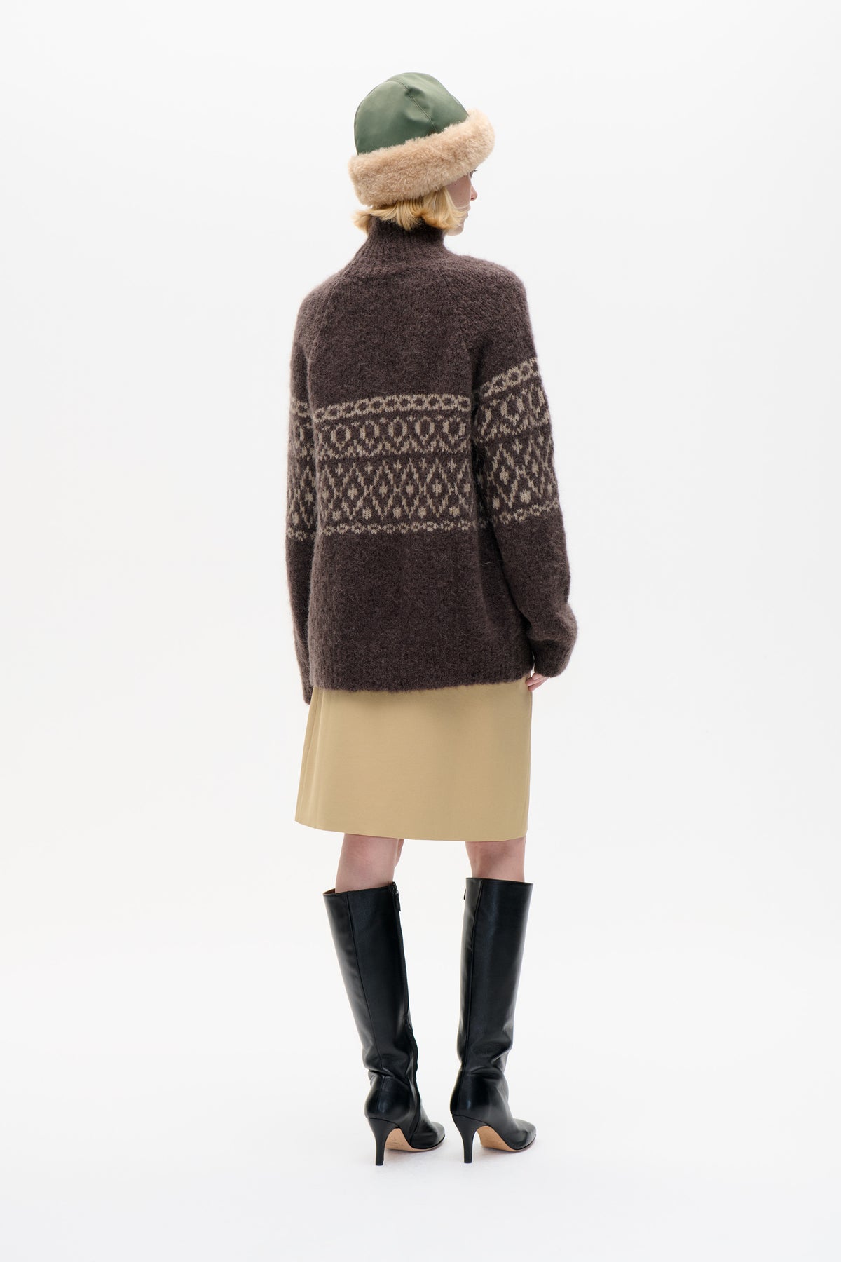 Rear three-quarter view of a model wearing a dark brown sweater with cream patterned bands, a green hat with a beige fuzzy brim, and a tan skirt.