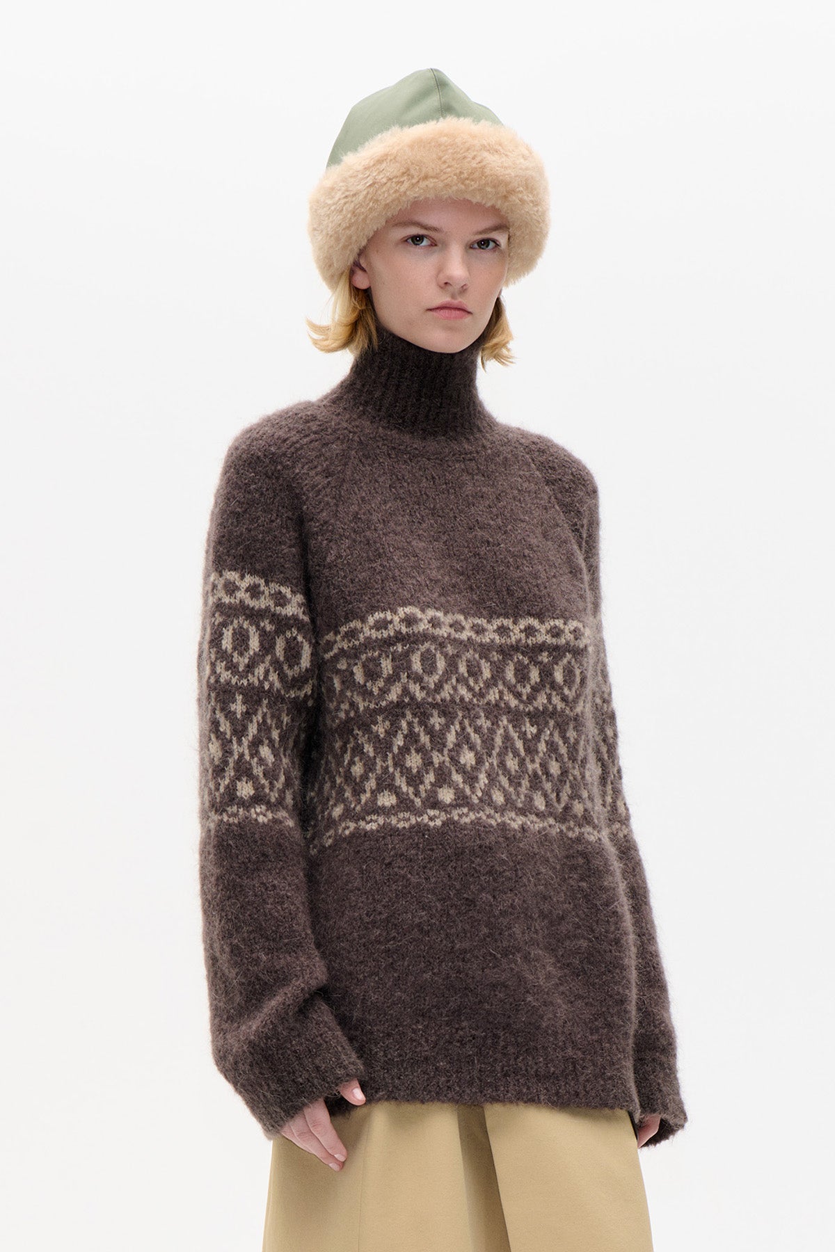 Model wearing a brown high-neck sweater with a cream geometric band across the chest and sleeves, paired with beige trousers.