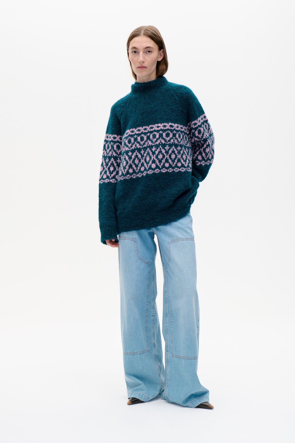 Model wearing a teal high-neck knit sweater with pink geometric patterned bands and light-wash wide-leg jeans.