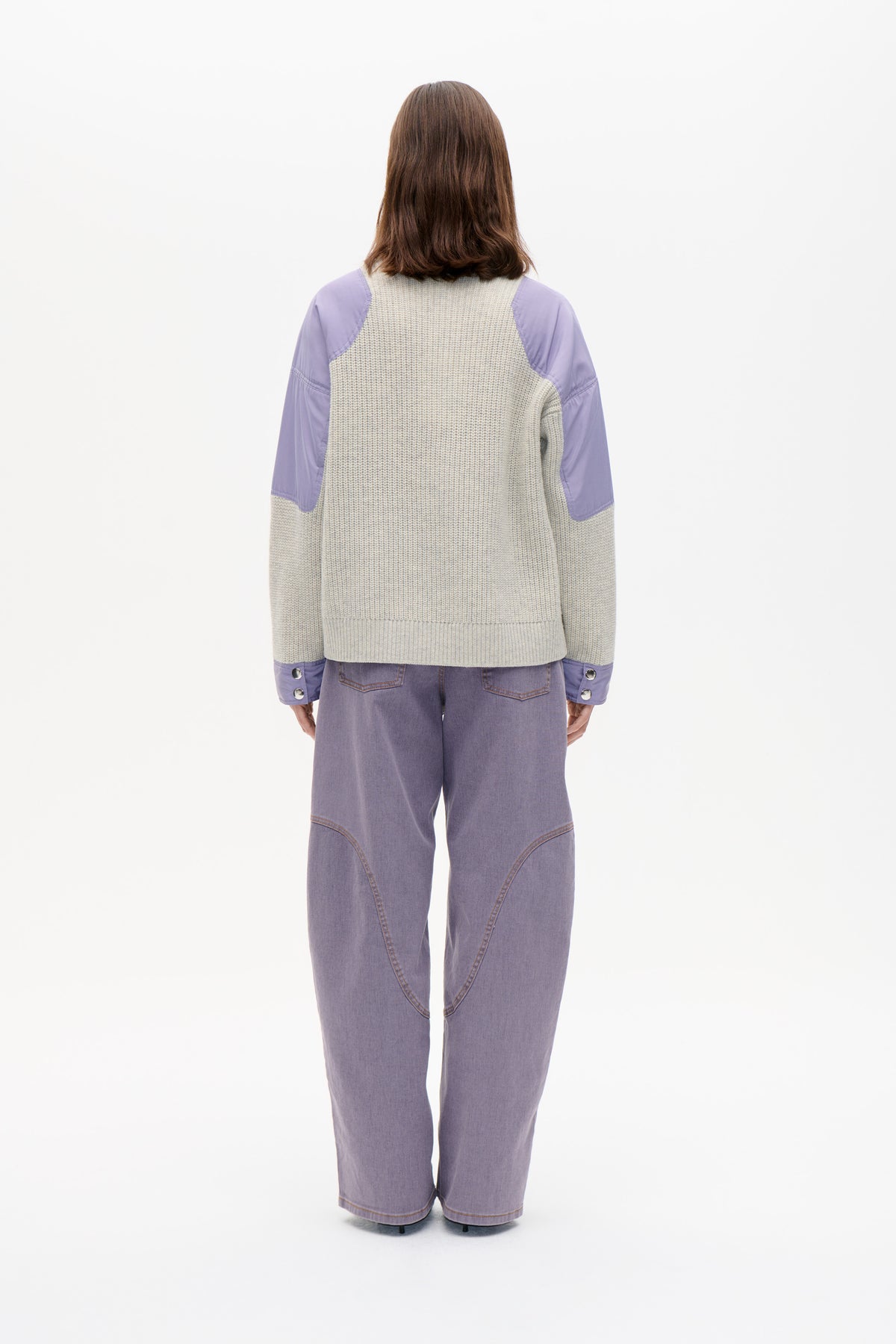 Back view of a chunky light grey knit sweater with lavender fabric panels on the shoulders and sleeves, worn with lavender jeans.