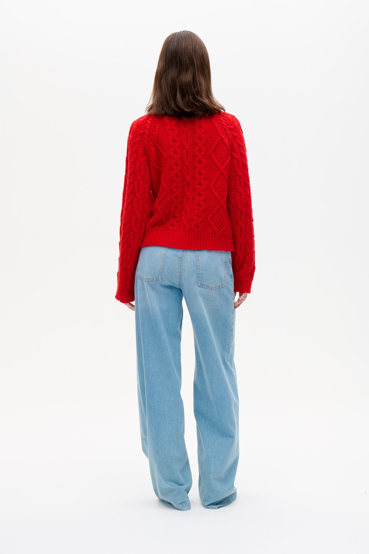 Back view of a model wearing a bright red cable-knit sweater with ribbed hem and light blue wide-leg jeans.