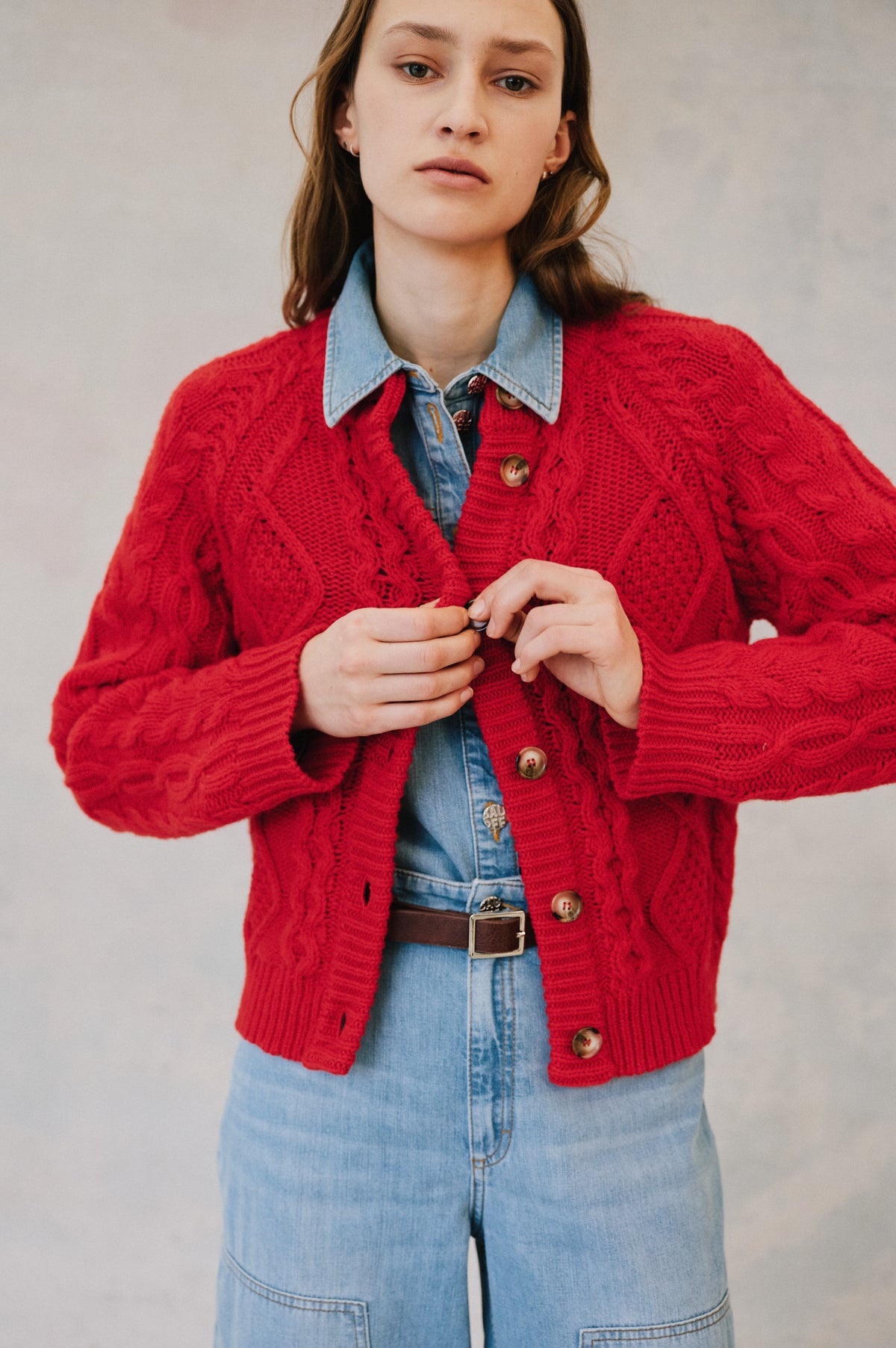 Campaign close-up of a model wearing a bright red cable-knit button cardigan over a light blue denim shirt and jeans.
