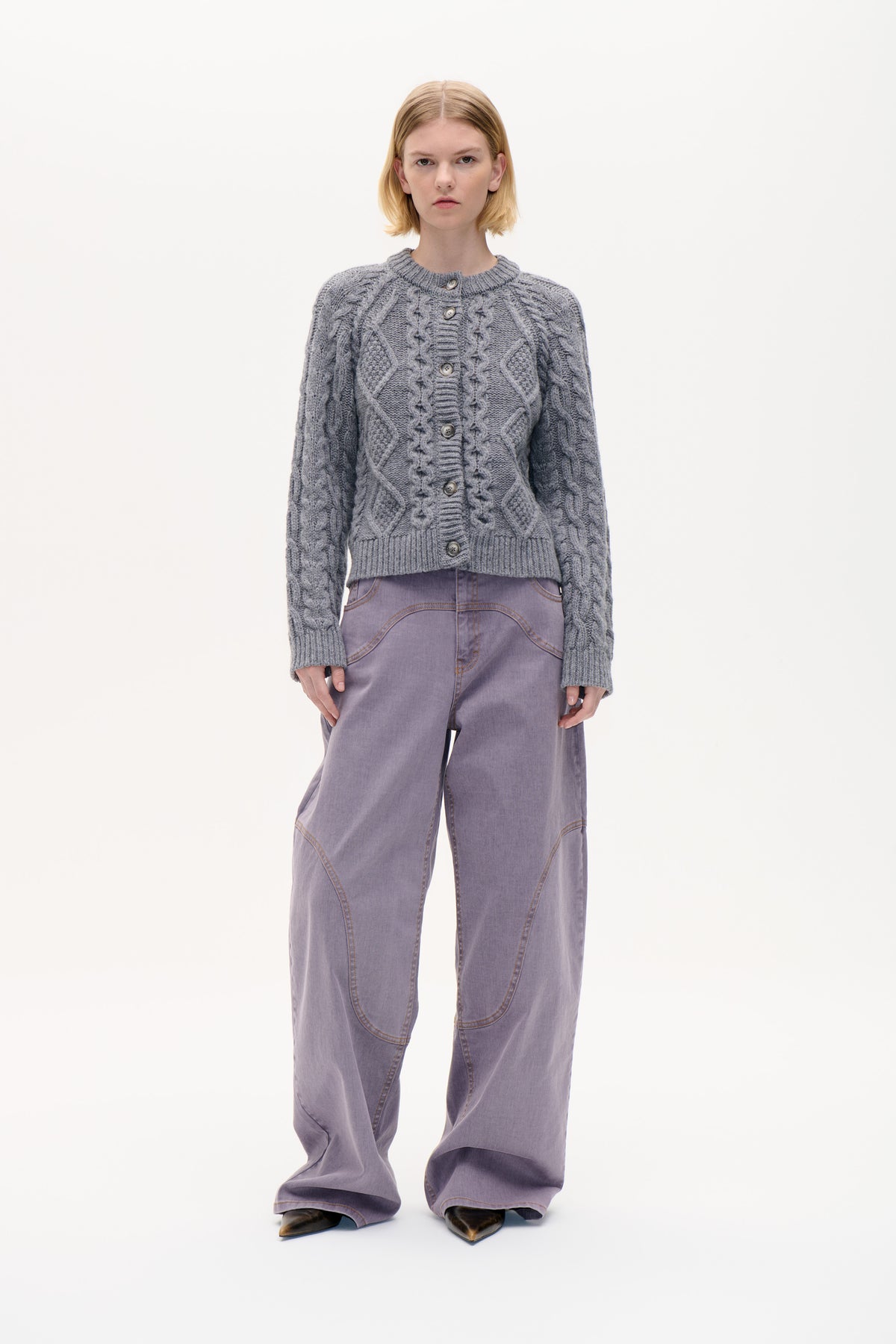 Front view of a model wearing a grey cable-knit button-front cardigan with ribbed trim and lavender wide-leg jeans.
