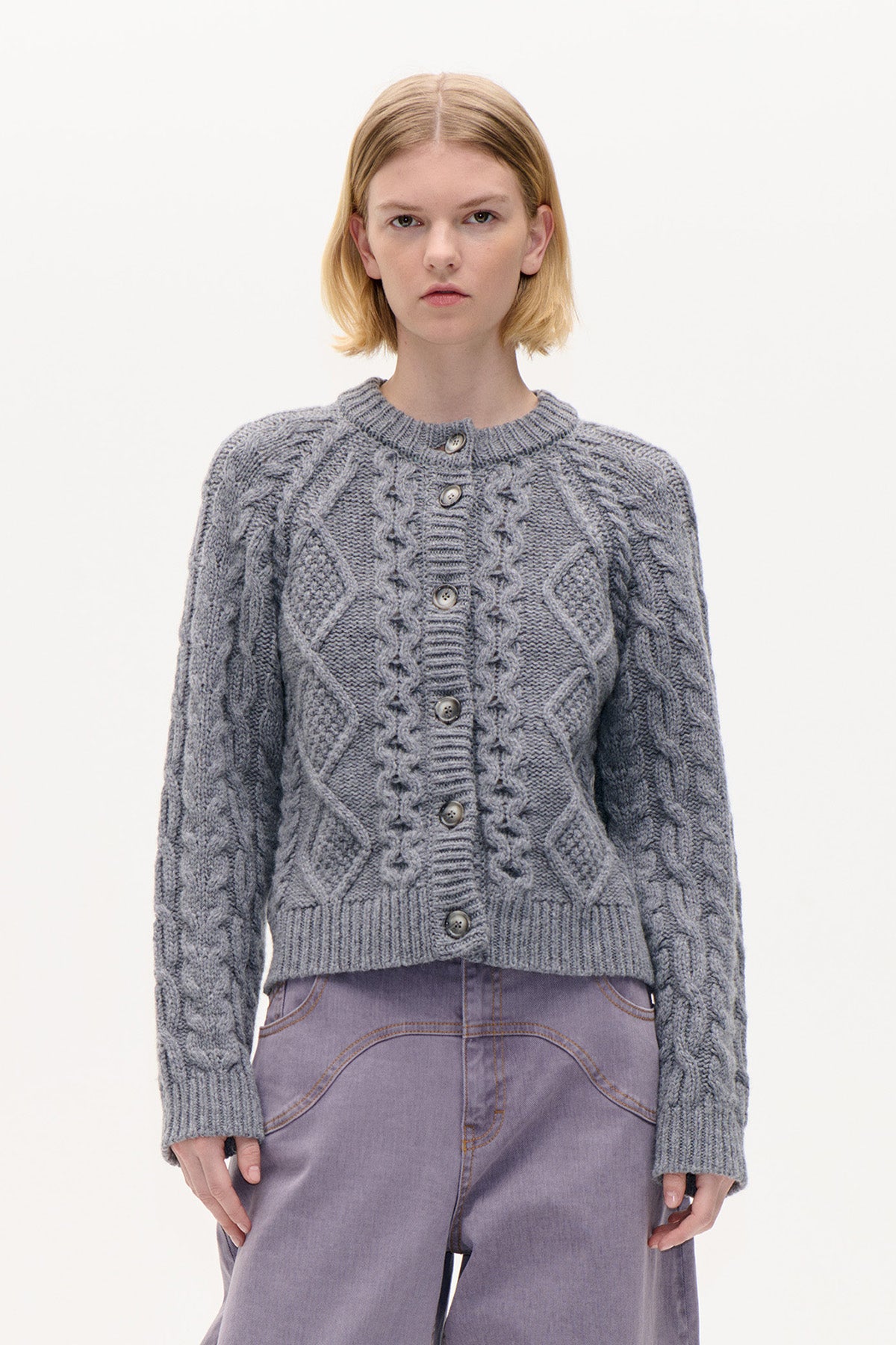 Front view of a grey cable-knit button cardigan with a crew neckline and ribbed trim, styled with lilac trousers.
