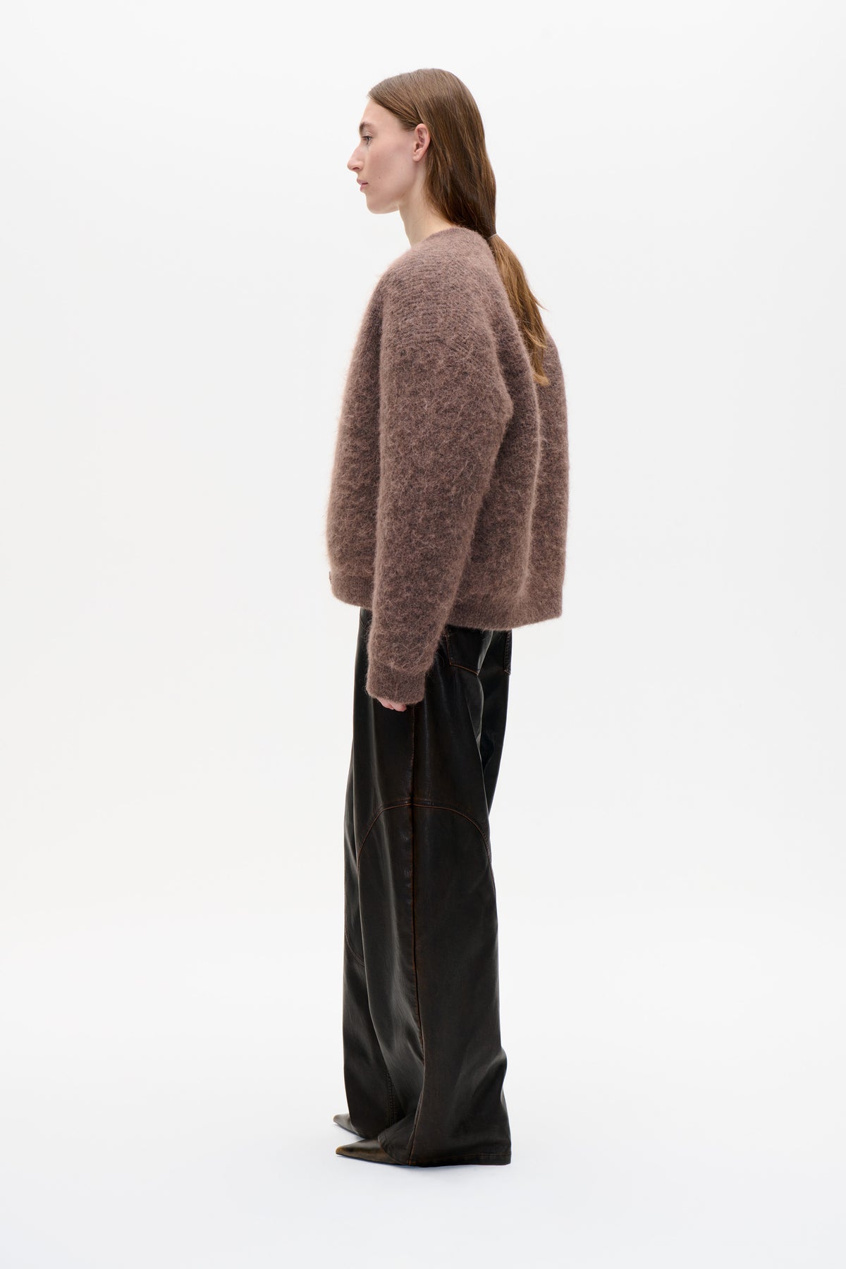 Side view of a model wearing a loose, fuzzy mauve-brown crewneck sweater with long sleeves and black wide-leg trousers.