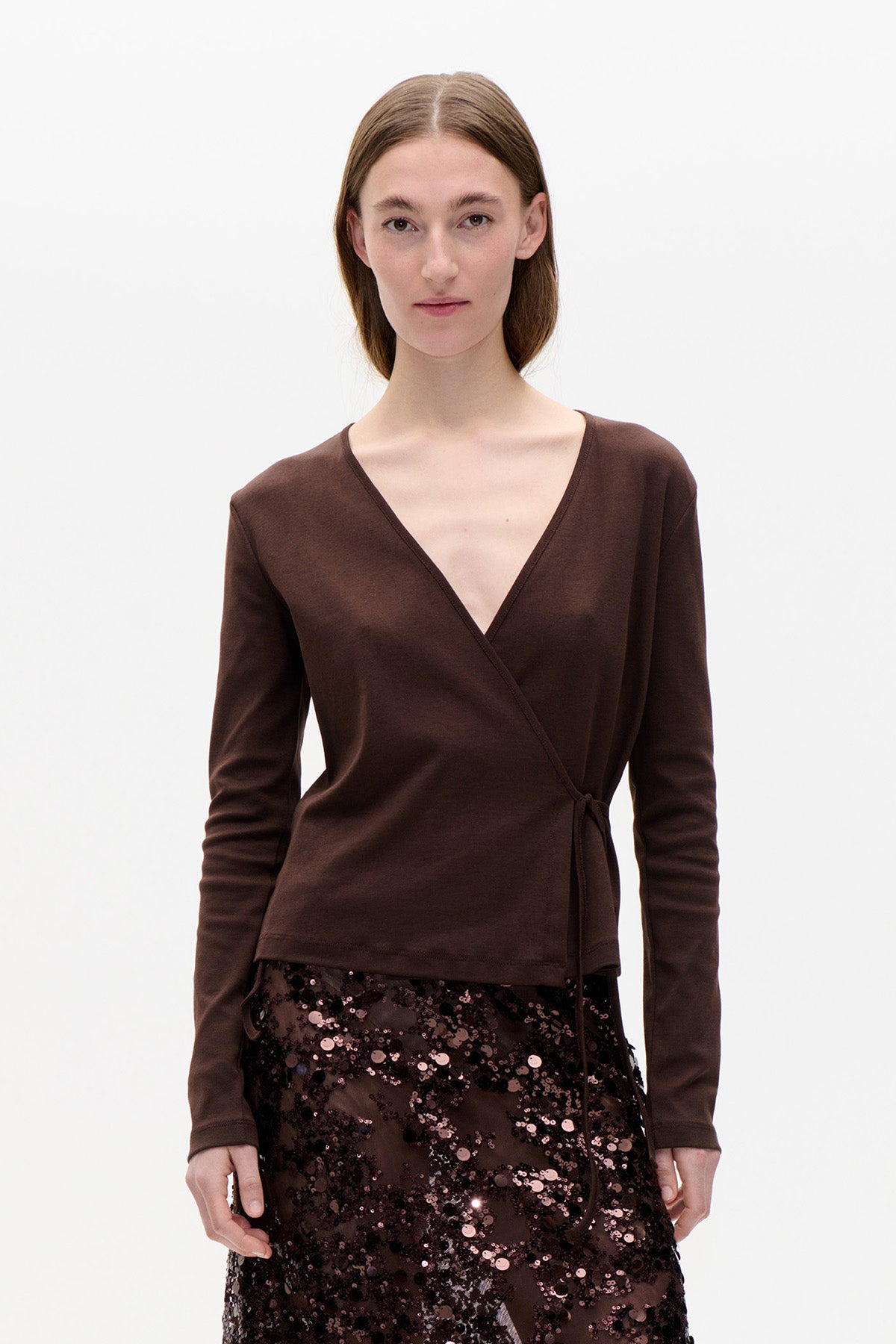 Model trägt ein dunkelbraunes Wickeltop mit langen Ärmeln, tiefem V‑Ausschnitt und Bindeband in der Taille, kombiniert mit einem transparenten Paillettenrock.