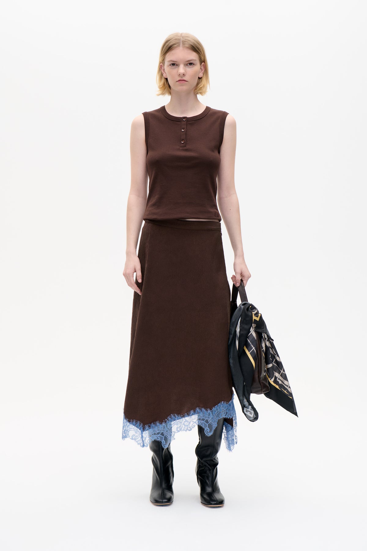 Front view of a model wearing a dark brown sleeveless button-front top and matching long skirt with pale blue lace trim, carrying a patterned bag.