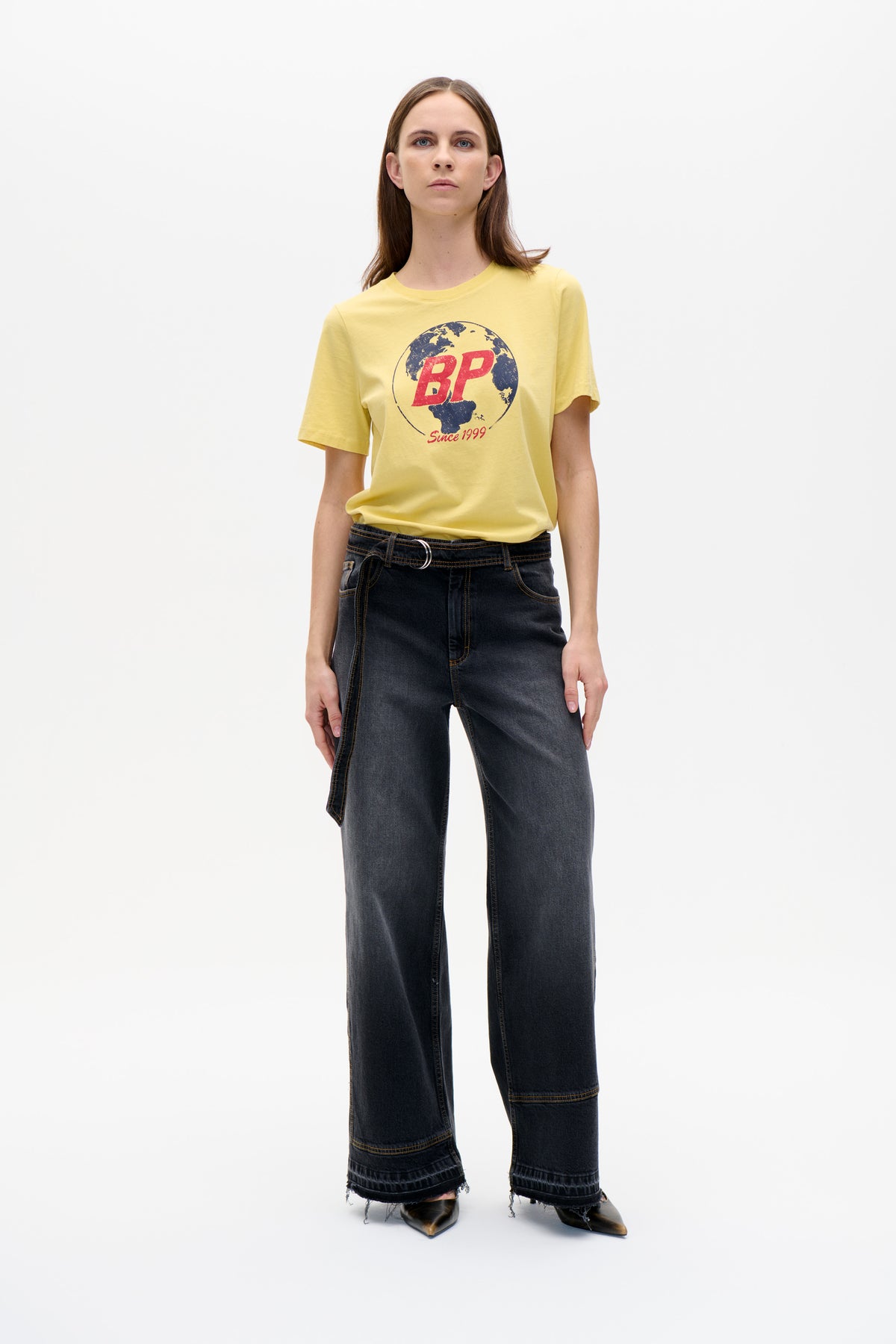 Front view of a model wearing a yellow short-sleeve T-shirt with a globe and red “BP Since 1999” graphic, paired with dark wide-leg jeans.