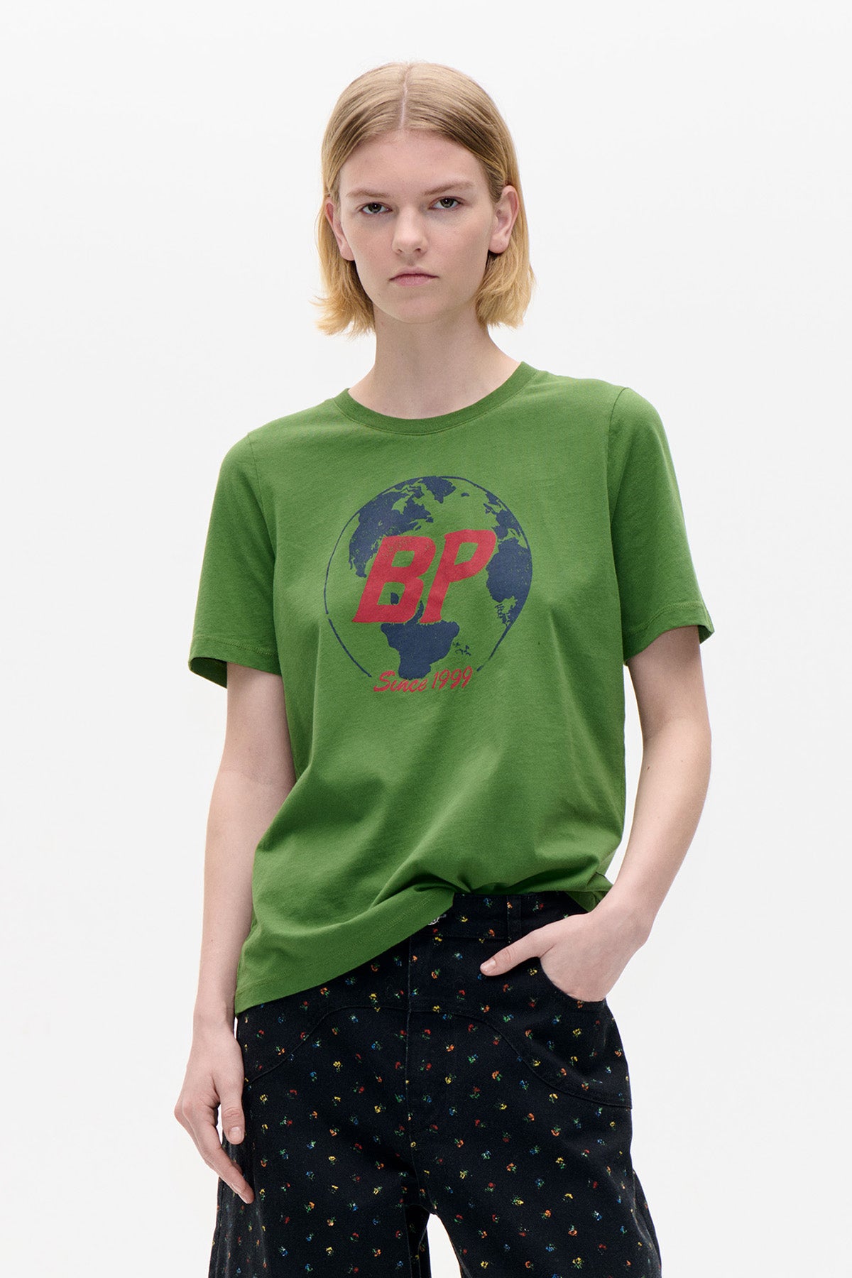 Front view of a green short-sleeved T-shirt with a navy globe and red “BP Since 1999” print, paired with black multicolour-speckled trousers.