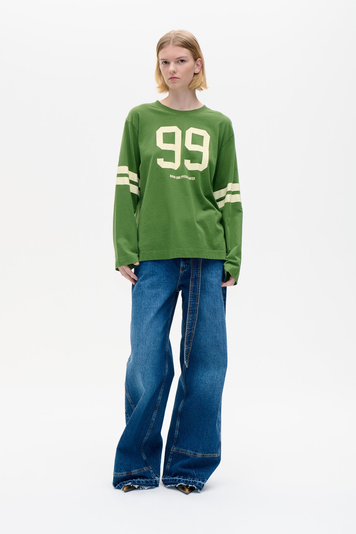 Model wearing a green long-sleeved sweatshirt with cream number 99 and striped sleeves, paired with wide-leg blue jeans.