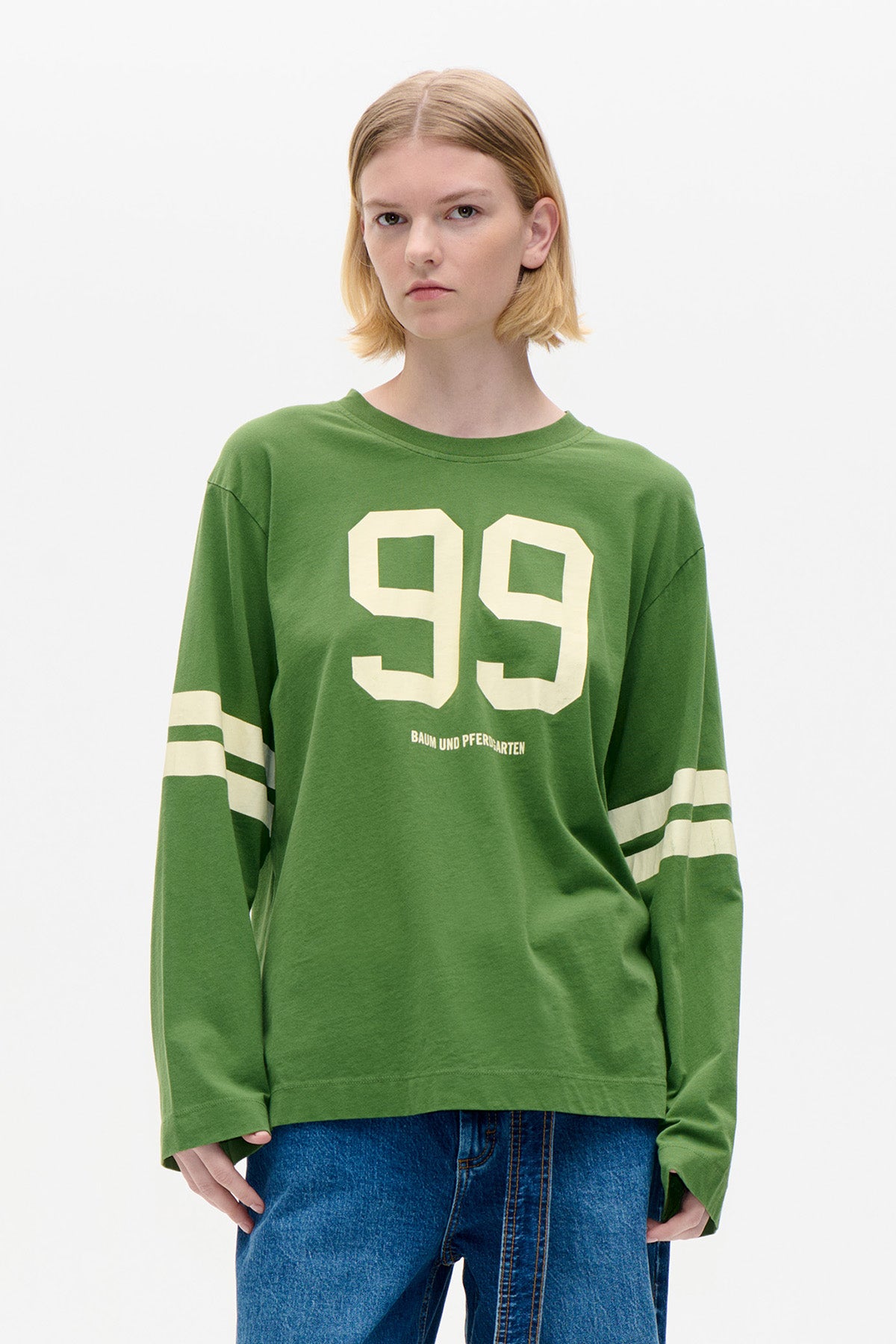 Front view of a model wearing a green long-sleeved T-shirt with cream number graphics and sleeve stripes, paired with blue jeans.
