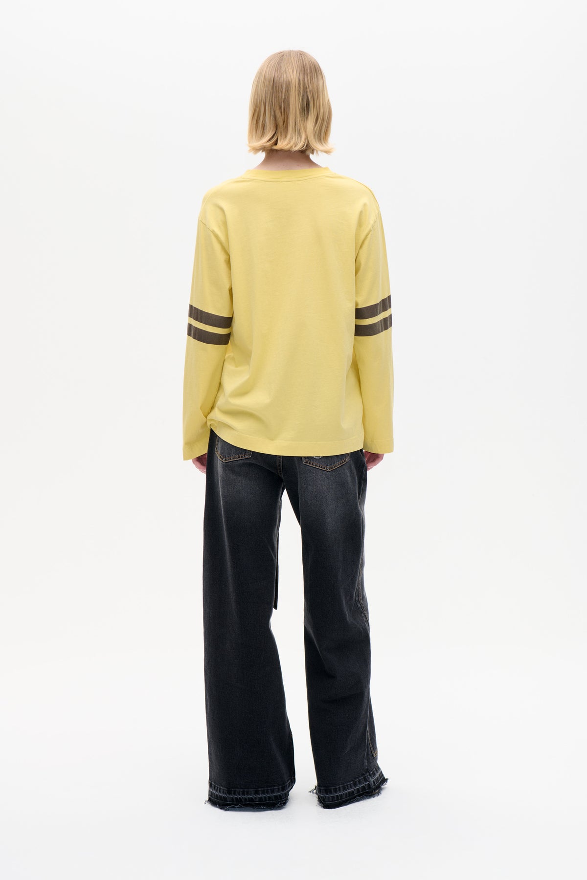 Back view of a model wearing a pale yellow long-sleeved top with dark stripe details across the sleeves and wide-leg black jeans.
