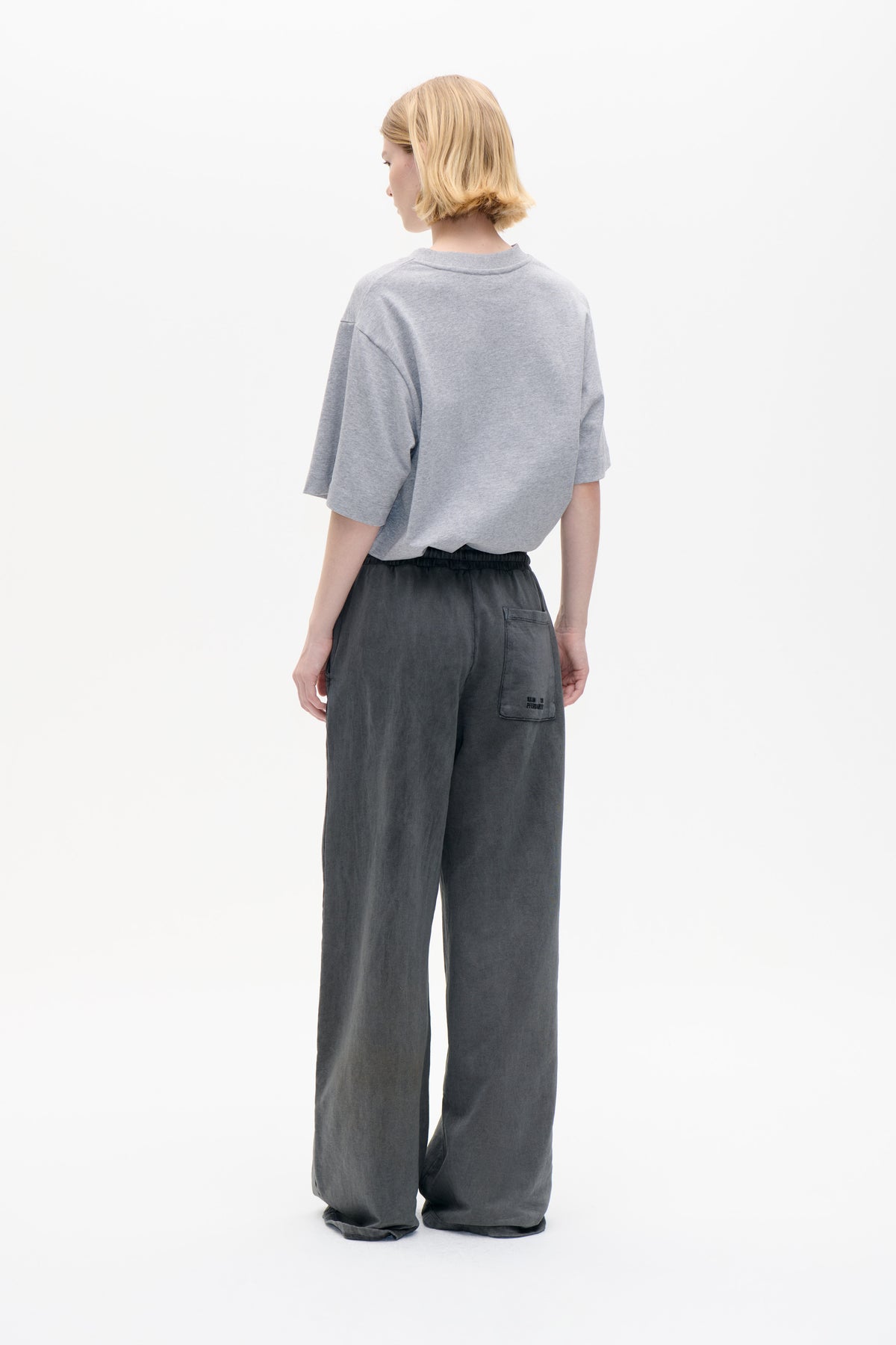 Back view of a model wearing a light grey oversized short-sleeved T-shirt and charcoal wide-leg trousers with an elastic waist and back pocket.